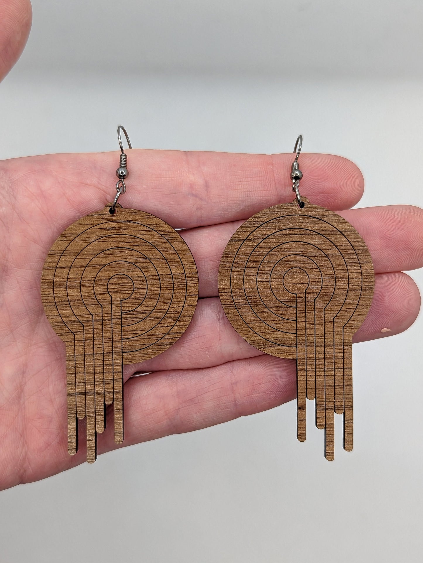 A pair of circle earrings with black ear wire hook fixings. Engraved into the centre is an 80s Retro melting record. They are made from walnut wood. They are against a hand to show the size of three finger widths in length and two wide