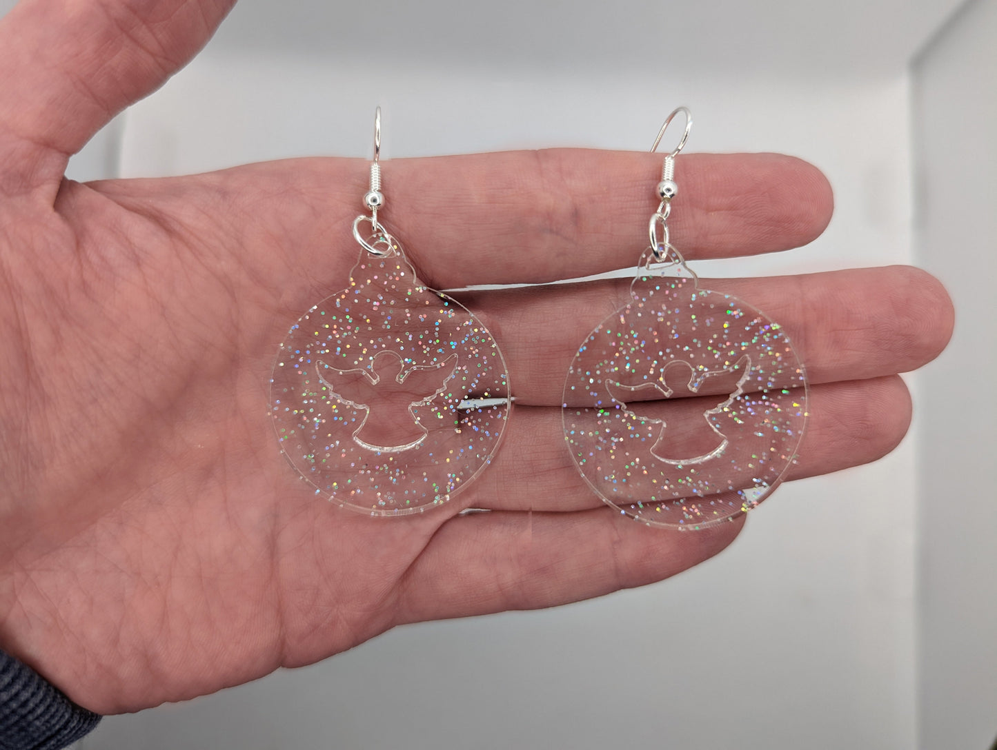 Image of a pair of earrings. They are Christmas / Xmas bauble shapes with an angel cut out in clear sparkle on a hand showing the size against a white background, they are 2 finger widths length the same again wide. They and have silver fixings.