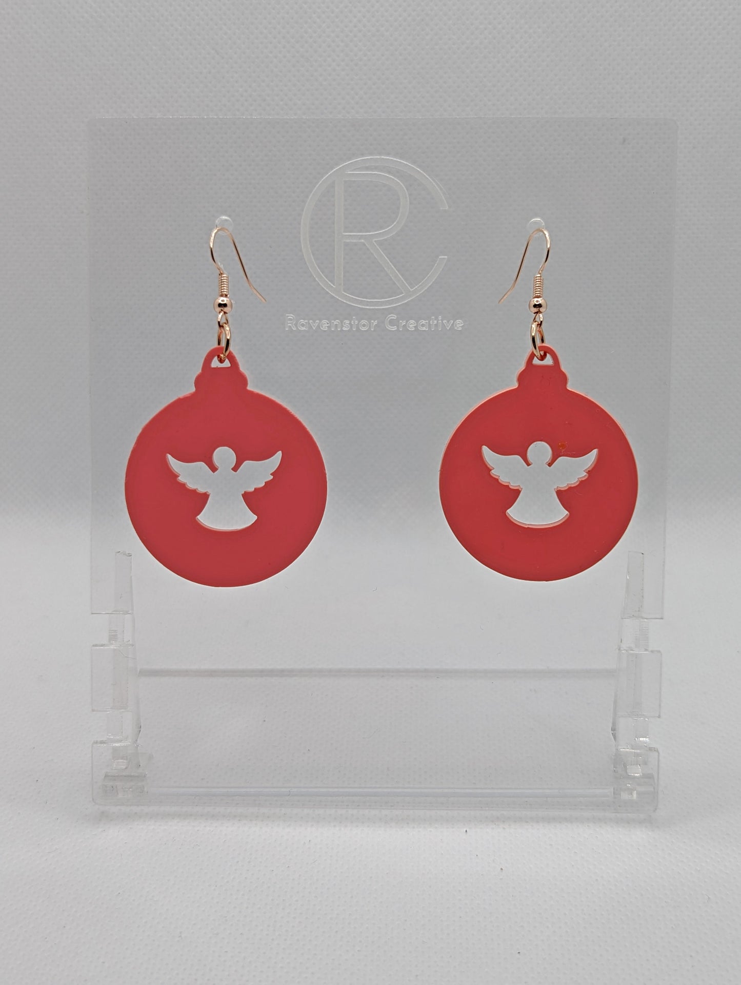 Shows a pair of acrylic earrings that have rose gold ear wire hook fixings. They are Christmas / Xmas baubles shapes with an angel shape cut out the middle. The colour is sherbet pink. They are against a white background on see through stands.