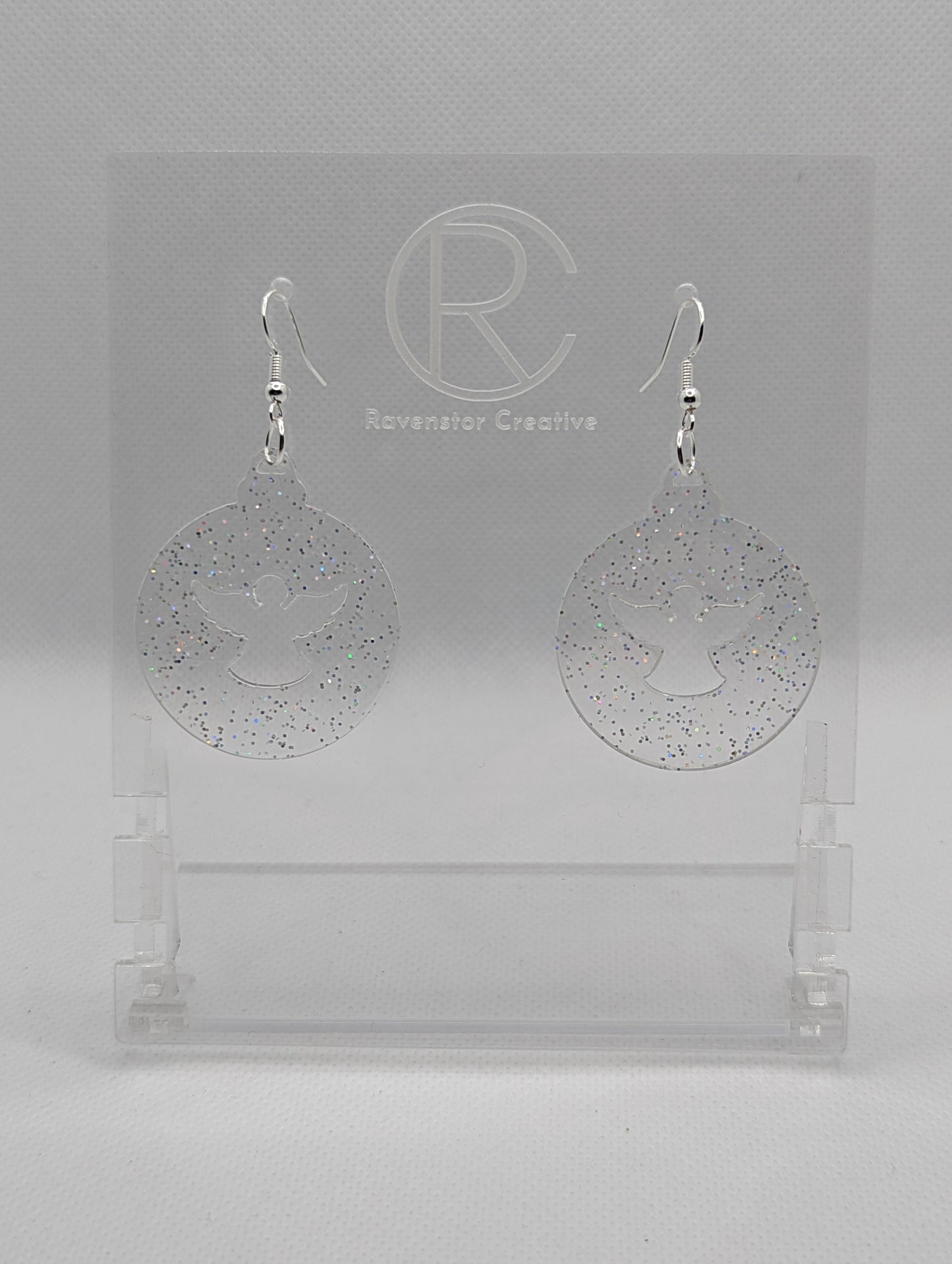 Shows a pair of acrylic earrings that have silver ear wire hook fixings. They are Christmas / Xmas baubles shapes with an angel shape cut out the middle. The colour is clear sparkle. They are against a white background on see through stands.