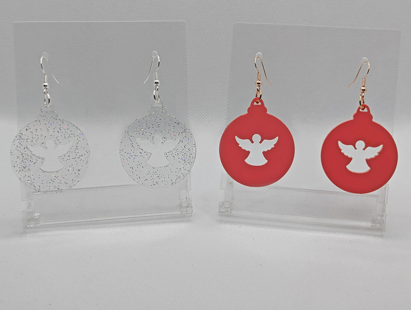 Shows two pairs of earrings that have ear wire hook fixings. They are Christmas baubles with an angel shape cut out the middle. The colours are Clear Sparkle and Sherbet Pink. They are against a white background on see through stands.