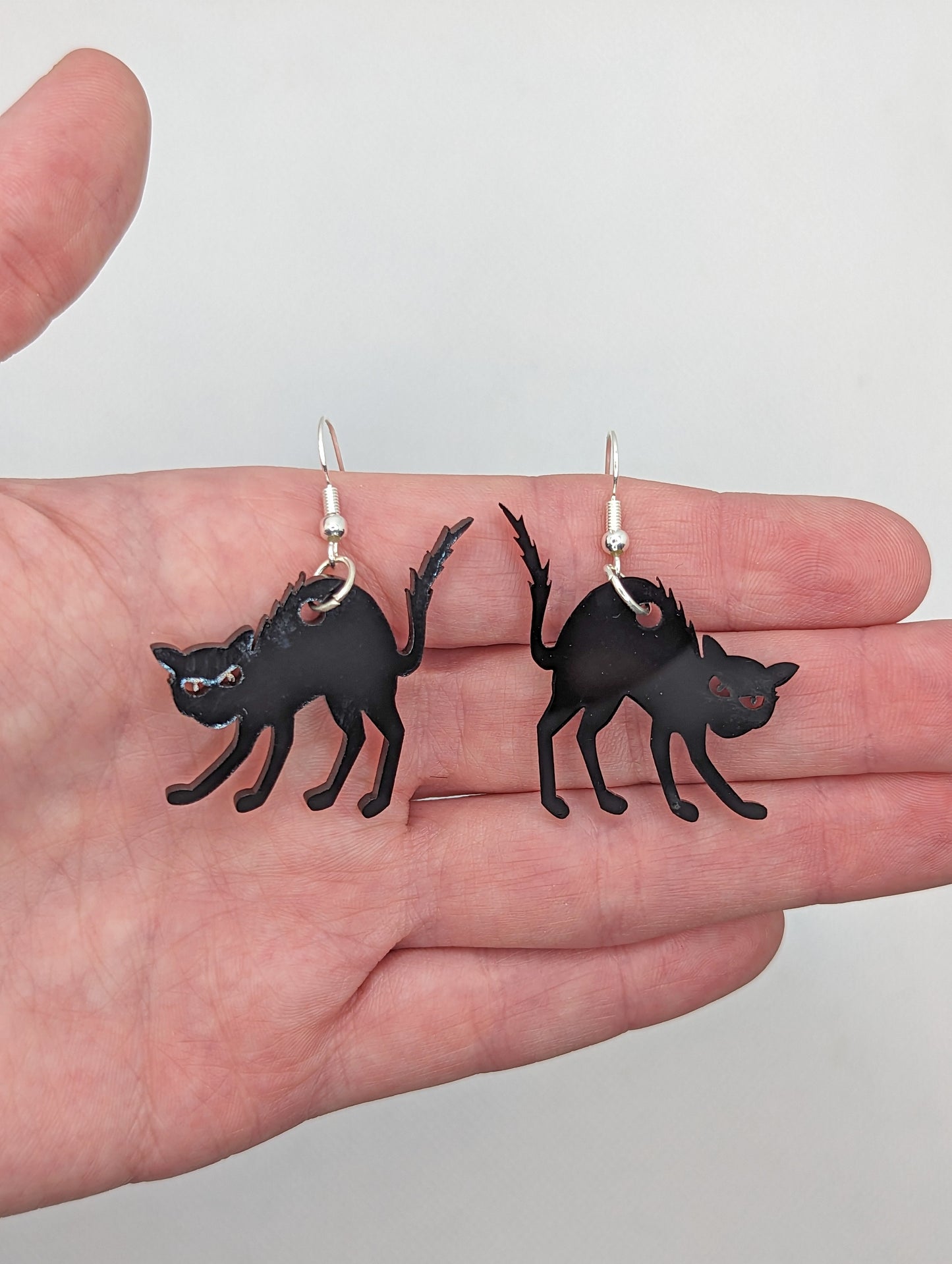 A pair of earrings in a hand to show the size with silver fixings, they are angry or scared looking cats in black with thier heckles and tails up and fir on end. They are black, the earrings themselves are about 2 fingers width and length