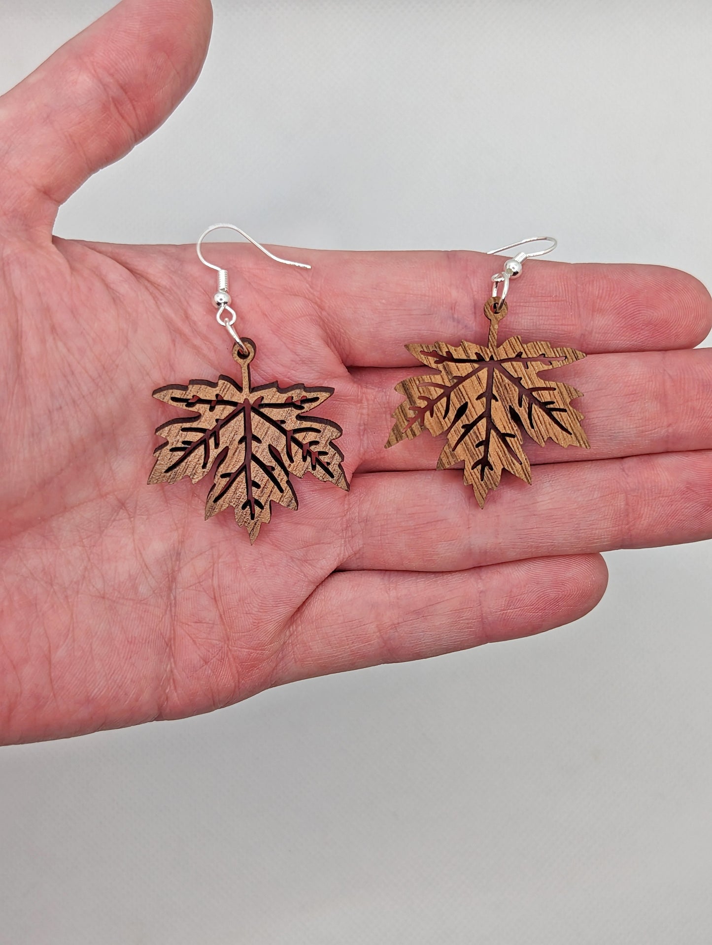 Image of a pair of Autumn Fall Maple Leaves on a hand showing the size against a white background, they are 1 and a half finger widths length the same again wide. They are walnut wood and have silver fixings.