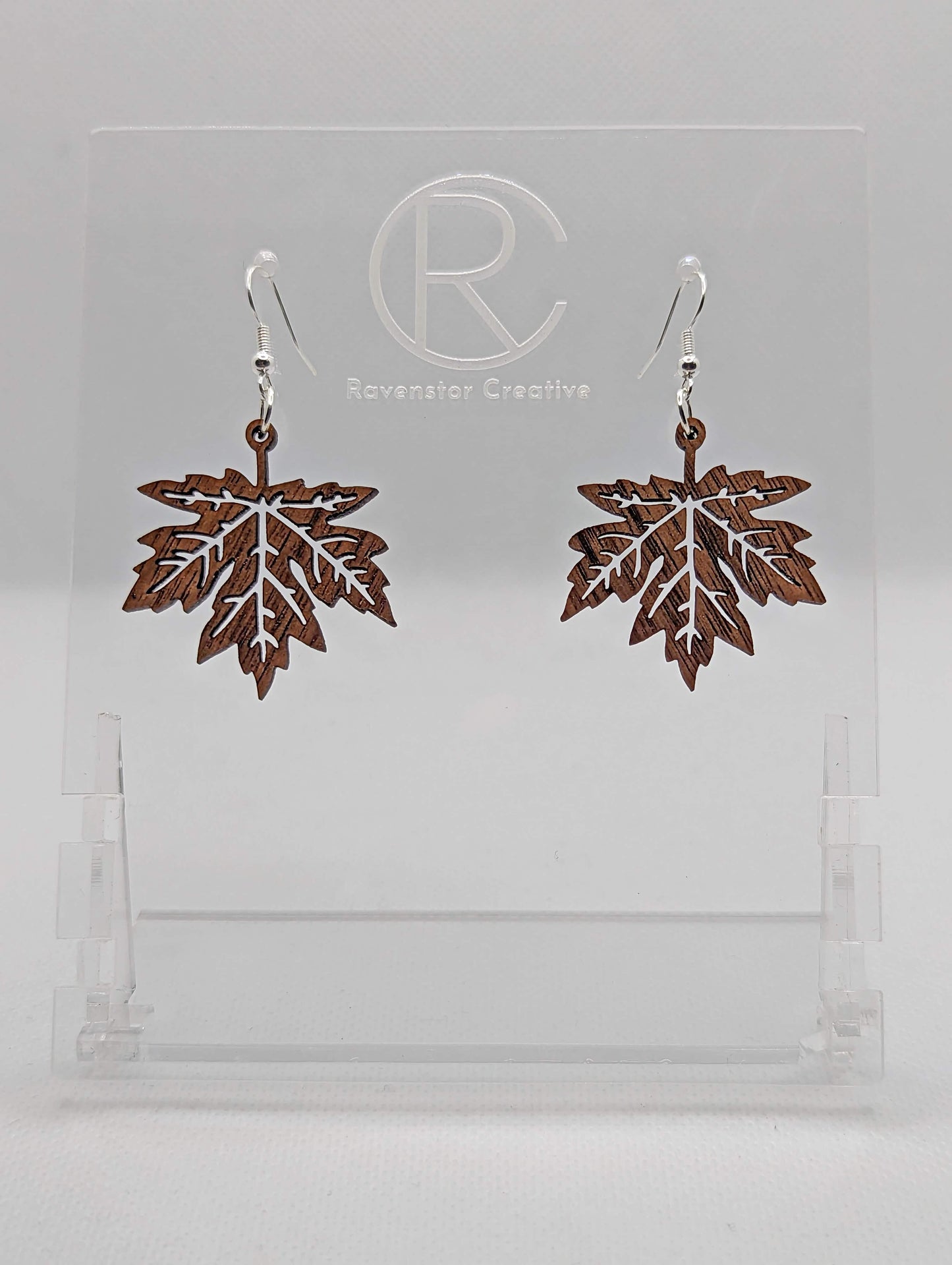 Shows a pair of earrings that have silver ear wire hook fixings. They are autumn fall maple leaf shapes. They are in Walnut wood and are against a white background on see through stand with the Ravenstor Creative logo on it.