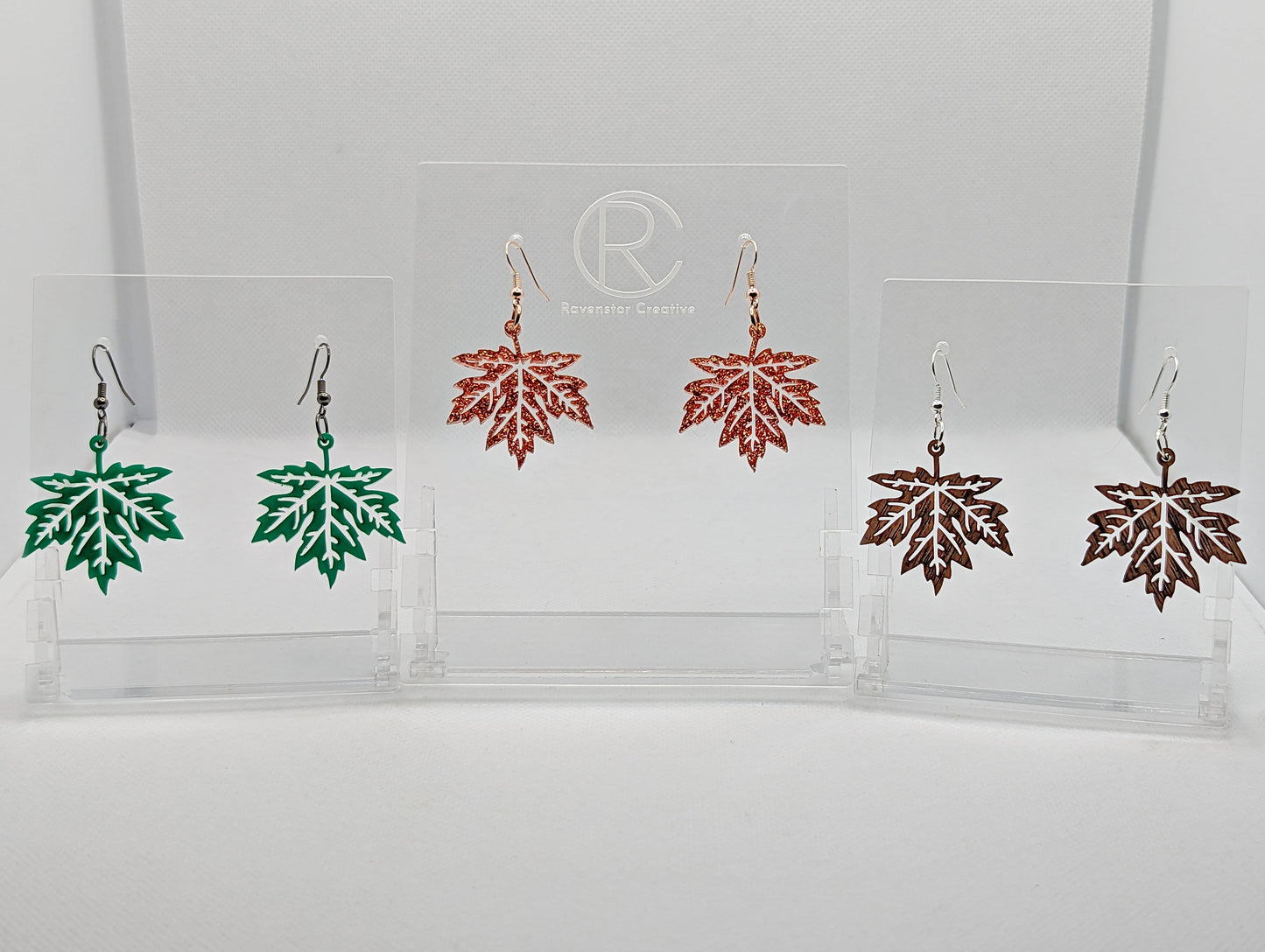Shows three pairs of earrings that have ear wire hook fixings. They are Autumn Fall Maple Leaf shapes. The colours are Green, Copper Glitter and Walnut Wood. They are against a white background on see through stands.