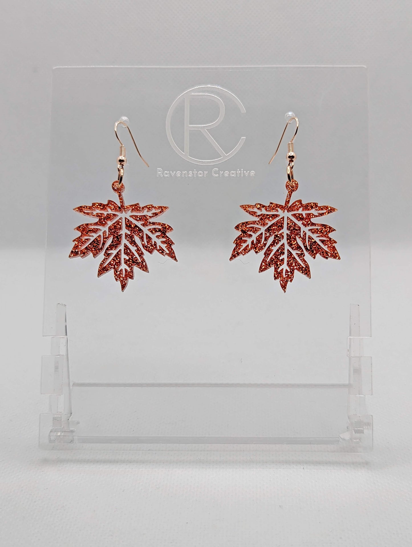 Shows a pair of acrylic earrings that have silver ear wire hook fixings. They are autumn fall maple leaf shapes. They are a copper glitter colour and are against a white background on see through stand with the Ravenstor Creative logo on it.