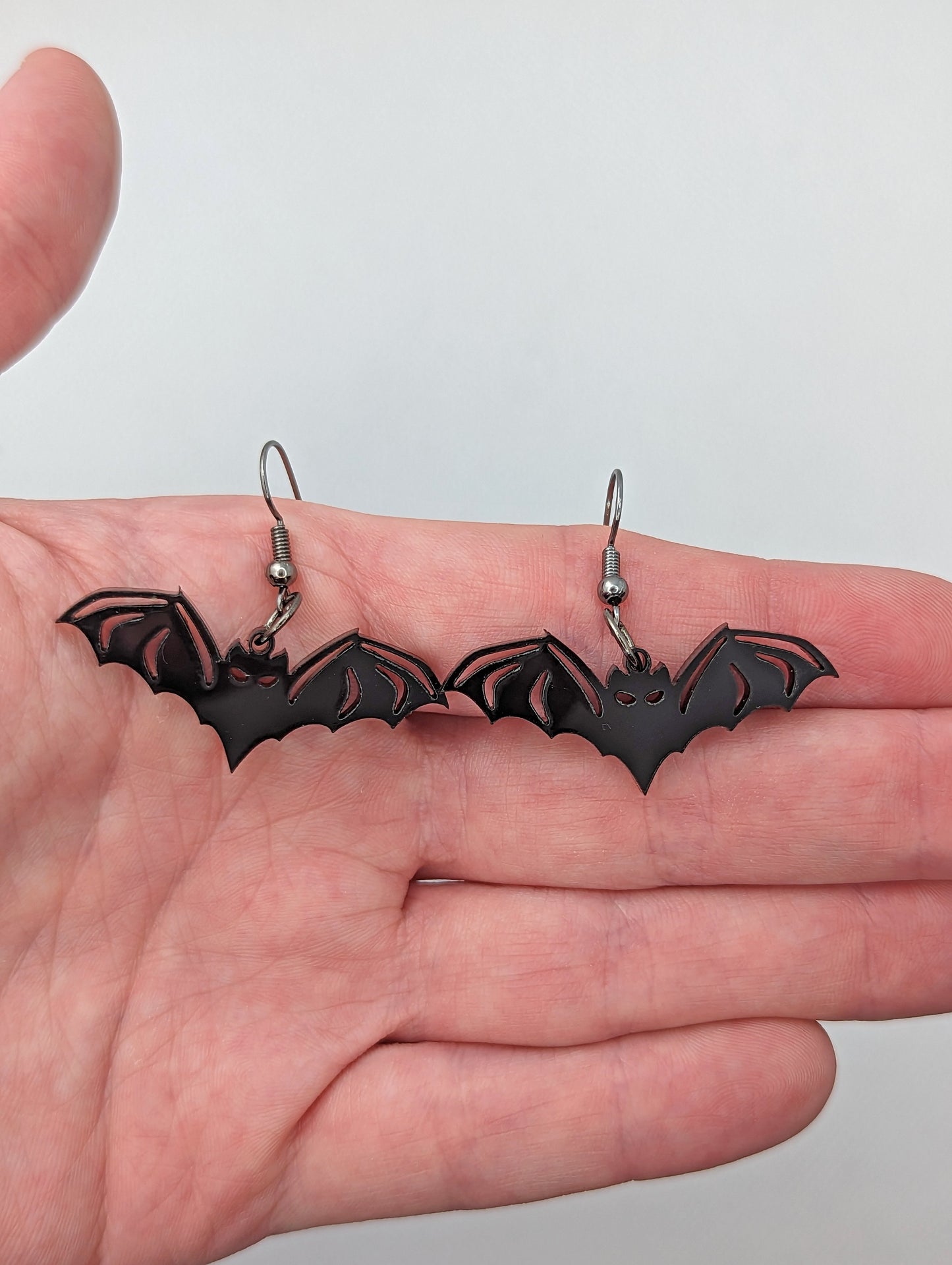 Image of a pair of Halloween Bat shaped earrings on a hand showing the size against a white background, they are one finger widths length and two wide. They are black and have black fixings.