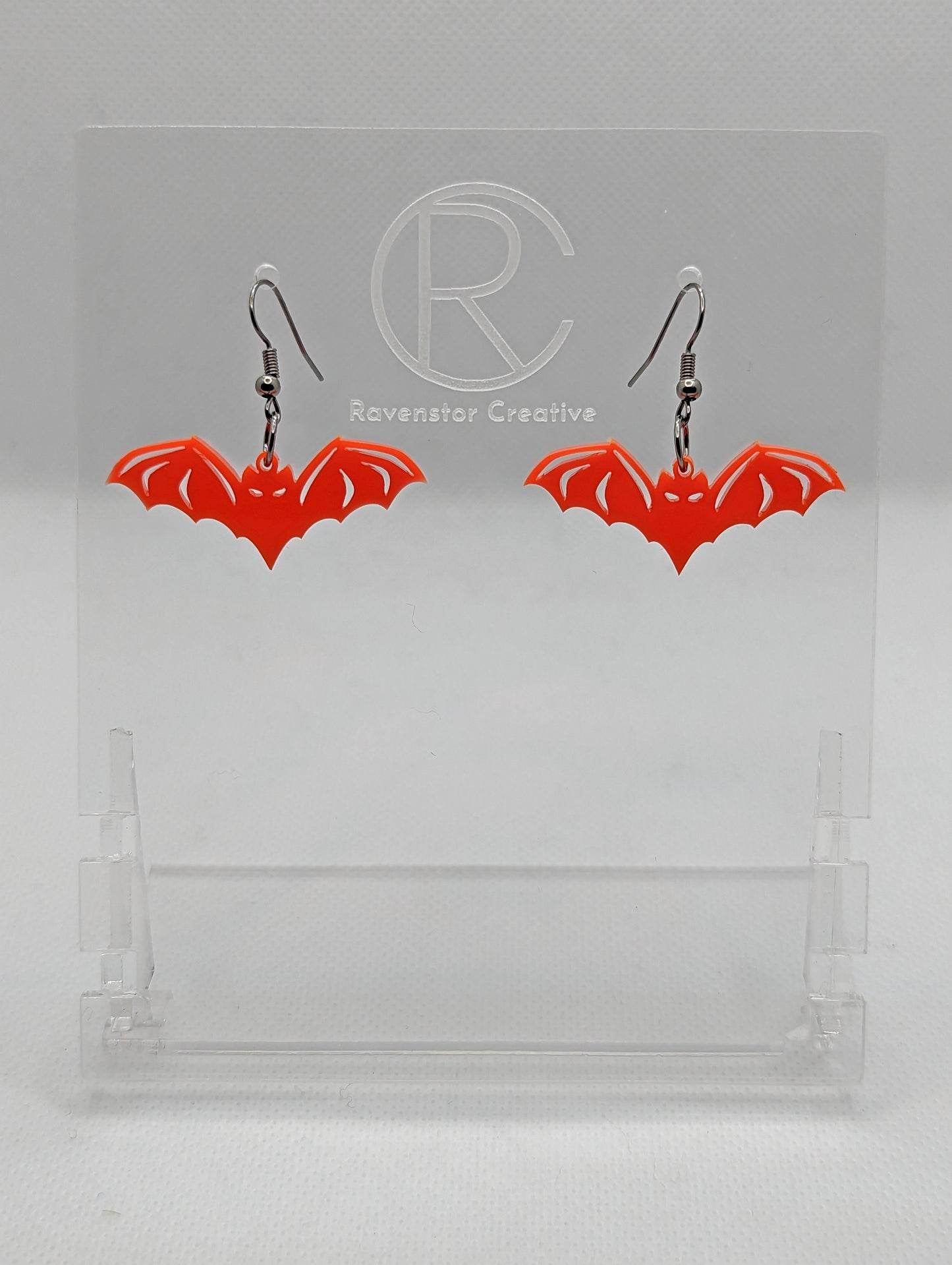 Shows a pair of earrings with black ear wire hook fixings. They are Halloween Bat shapes. The colour of them is red. They are on a see through stand that has the Ravenstor Creative Logo and its against a white background.