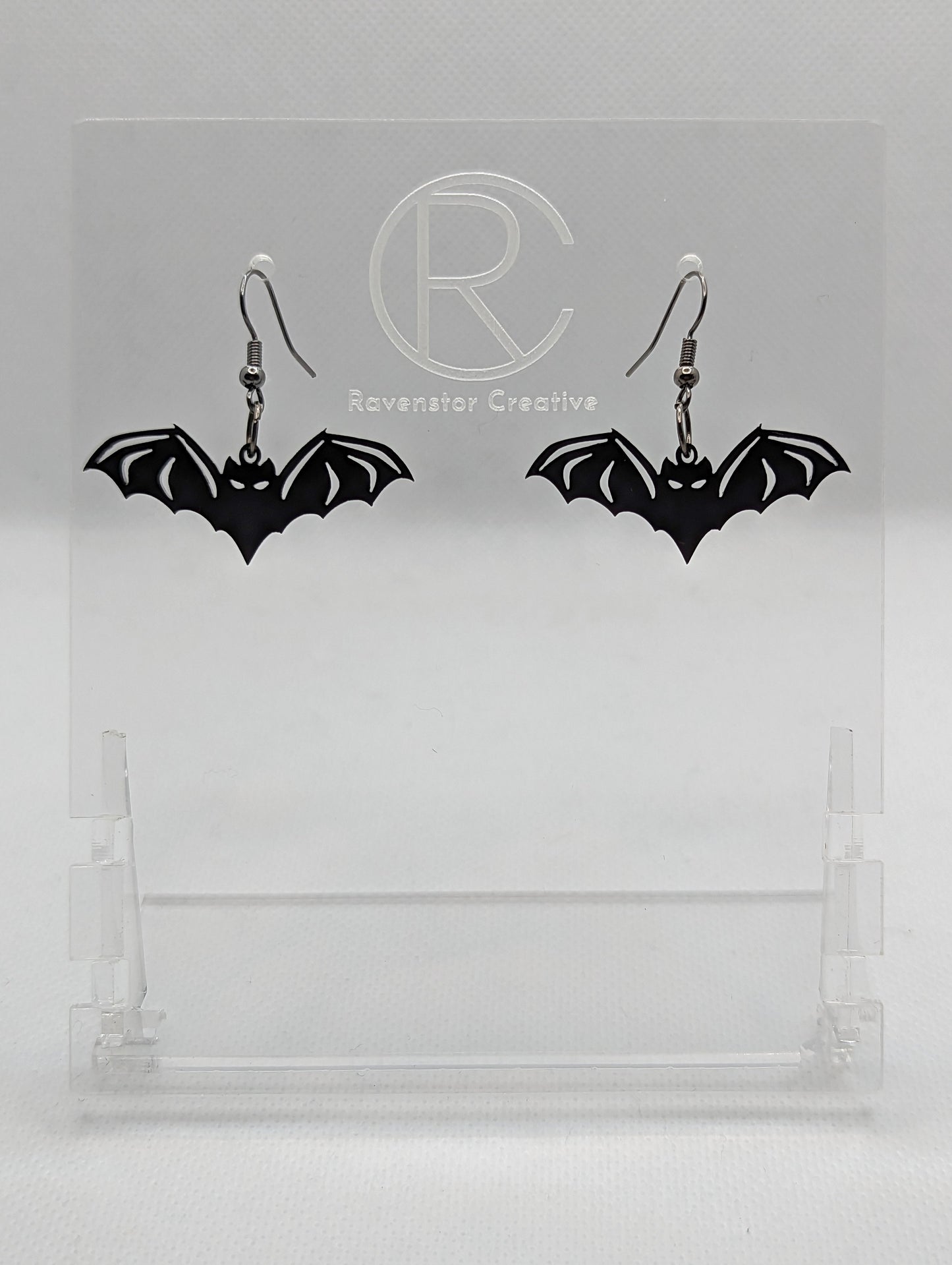 Image of a pair of Halloween Bat shaped earrings on a hand showing the size against a white background, they are one finger widths length and two wide. They are black and have black fixings.