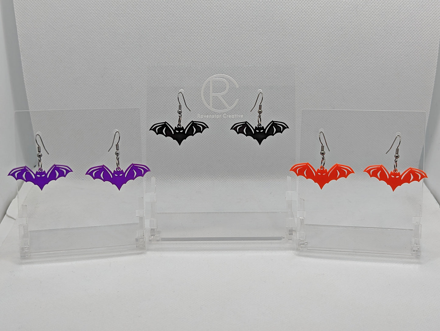 Shows 3 pairs of earrings with black ear wire hook fixings. They are Halloween Bat shapes. The colours of them are purple, black and red. They are on see through stands with the Ravenstor Creative logo on them against a white background