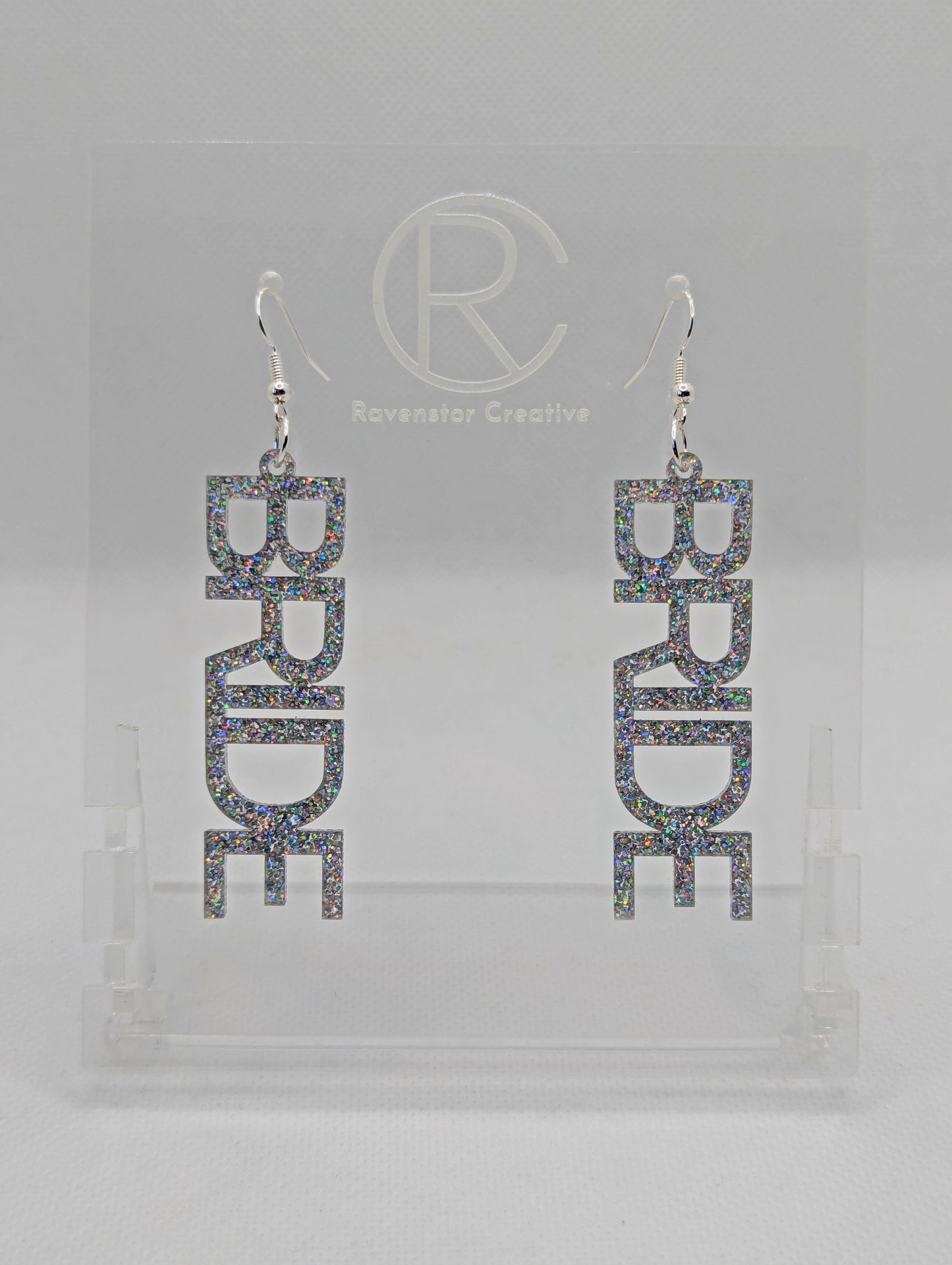 A pair of earrings with Silver ear wire hook fixings. Each one is the word Bride in capital letters. They are Holographic Glitter in colour. These are ideal for a Hen do or Bachelorette Party. They are on clear stands against a white background.