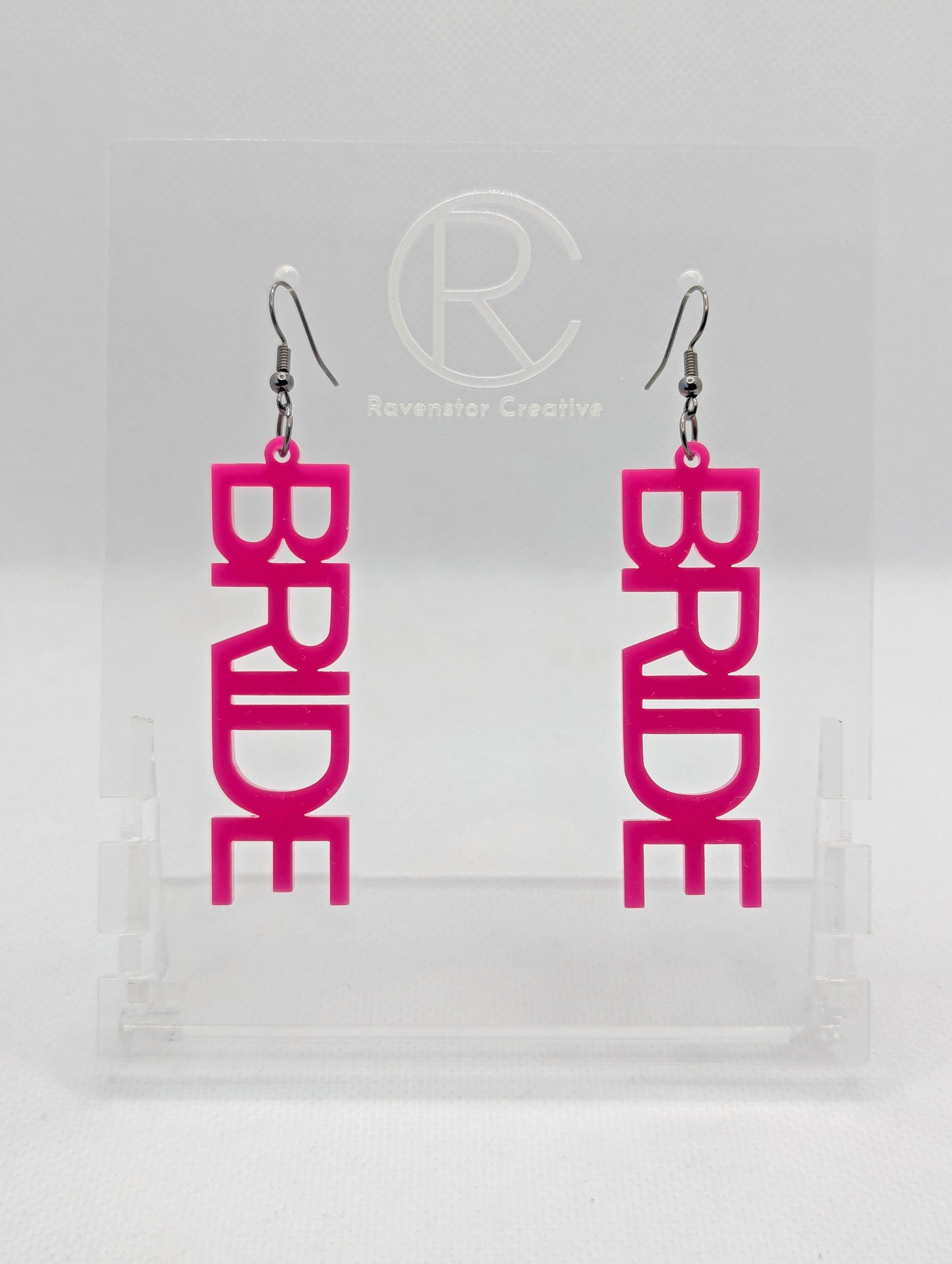 A pair of earrings with Black ear wire hook fixings. Each one is the word Bride in capital letters. They are Hot Pink in colour. These are ideal for a Hen do or Bachelorette Party. They are on clear stands against a white background.