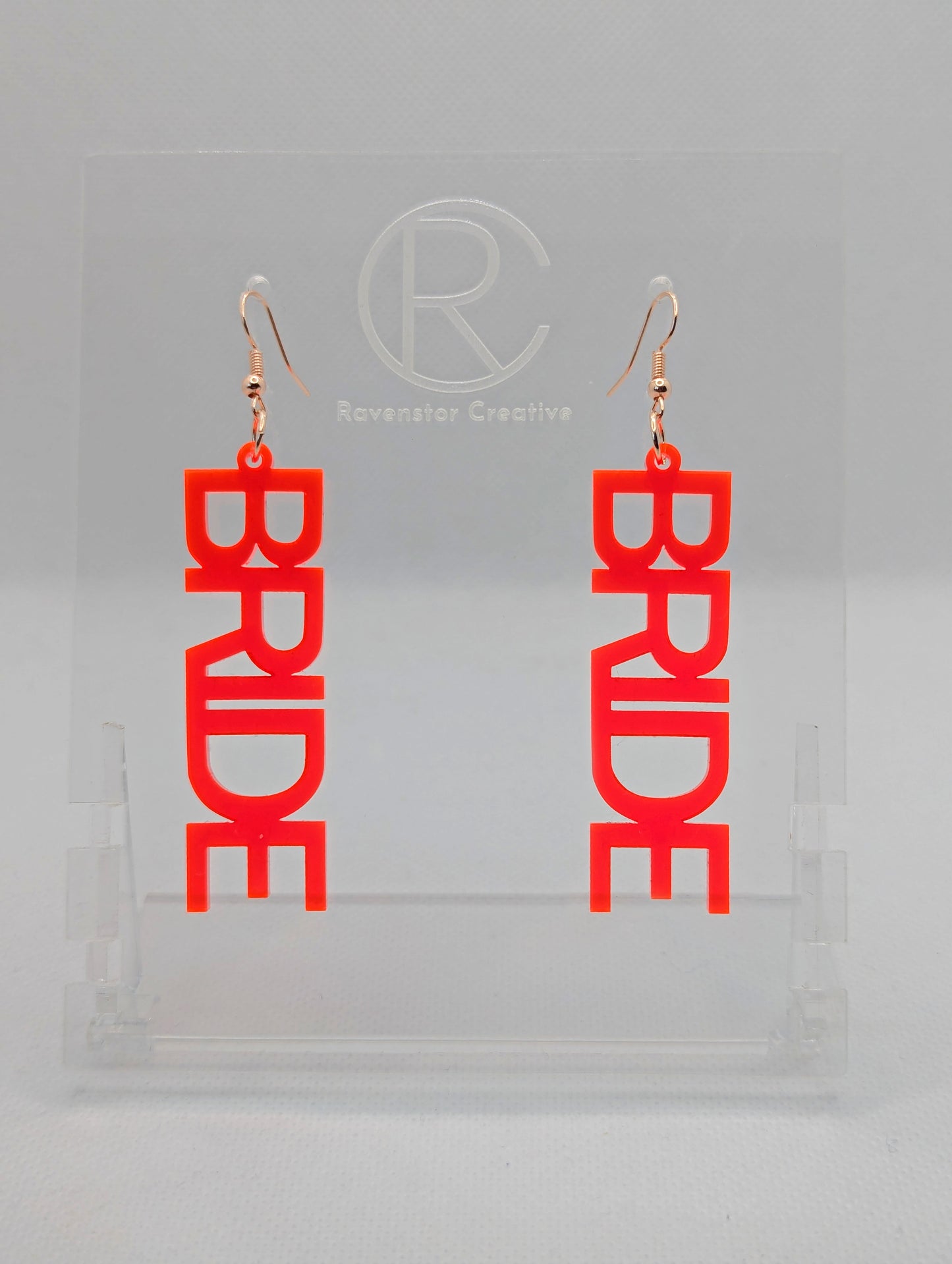 A pair of earrings with Rose Gold ear wire hook fixings. Each one is the word Bride in capital letters. They are Neon Pink in colour. These are ideal for a Hen do or Bachelorette Party. They are on clear stands against a white background.