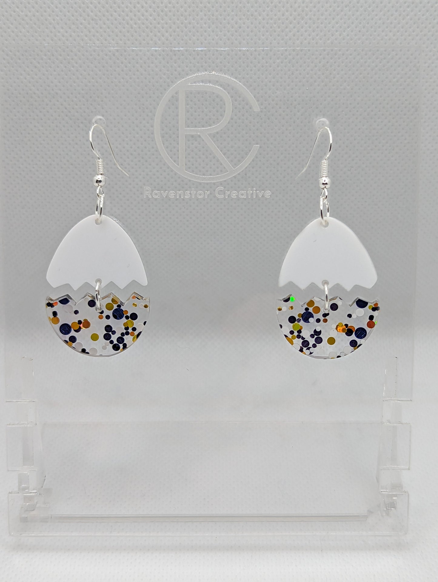 Shows a pair of Easter egg earrings cracked in the middle separating it into two parts. They have Silver ear wire hook fixings that attach to the top and the bottom dangles under. The colours are White top and Lollipop Glitter bottom.