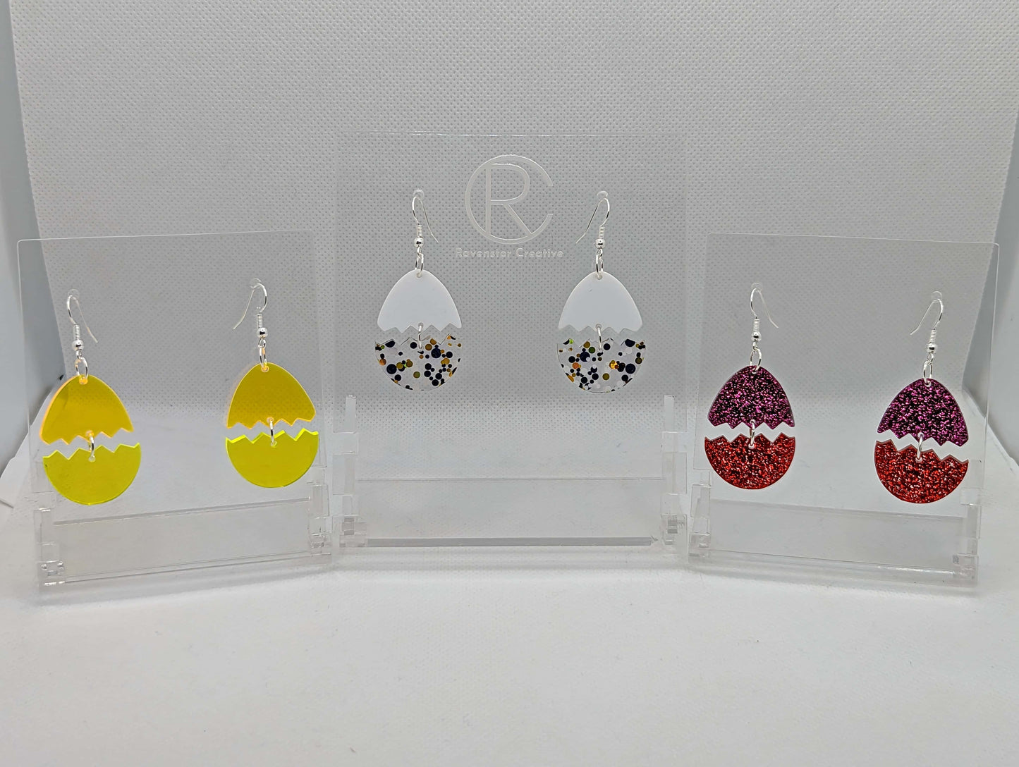 Shows 3 pairs of Easter egg earrings cracked in the middle separating it into two parts. Colours are Neon Orange top and neon yellow bottom, White top with Lollipop Glitter Bottom, Pink Glitter top with Red glitter bottom. They have ear wire fixings