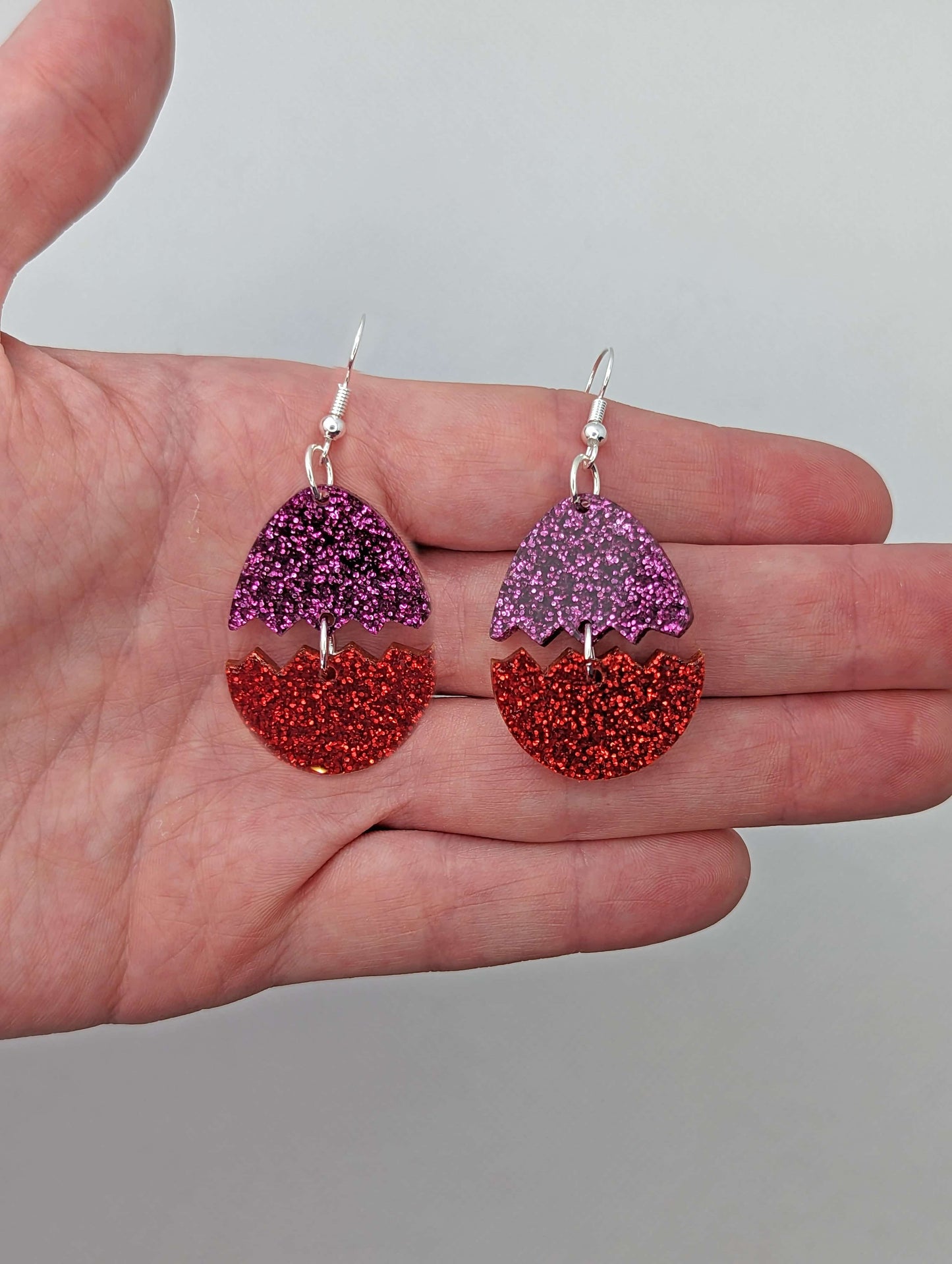 A pair of Easter egg earrings cracked in the middle separating it into two parts. They are against a hand showing the size of two finger widths long and two wide. They have ear wire hook fixings that attach to the top and the bottom dangles under.