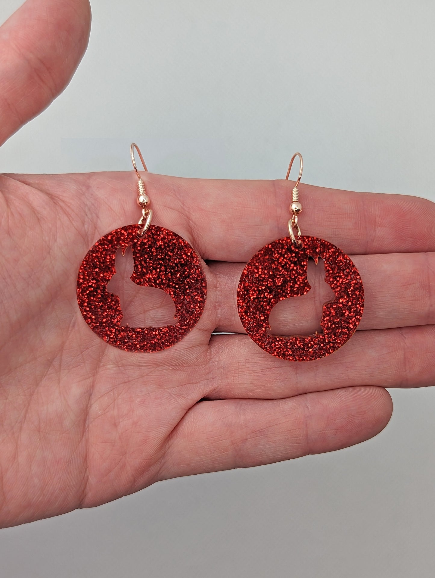 A pair of acrylic earrings that have Silver ear wire hook fixings against a hand to show the size of two finger widths long and same again wide. They are Circles with an Easter bunny / Rabbit cut out each one. It is Red Glitter colour.