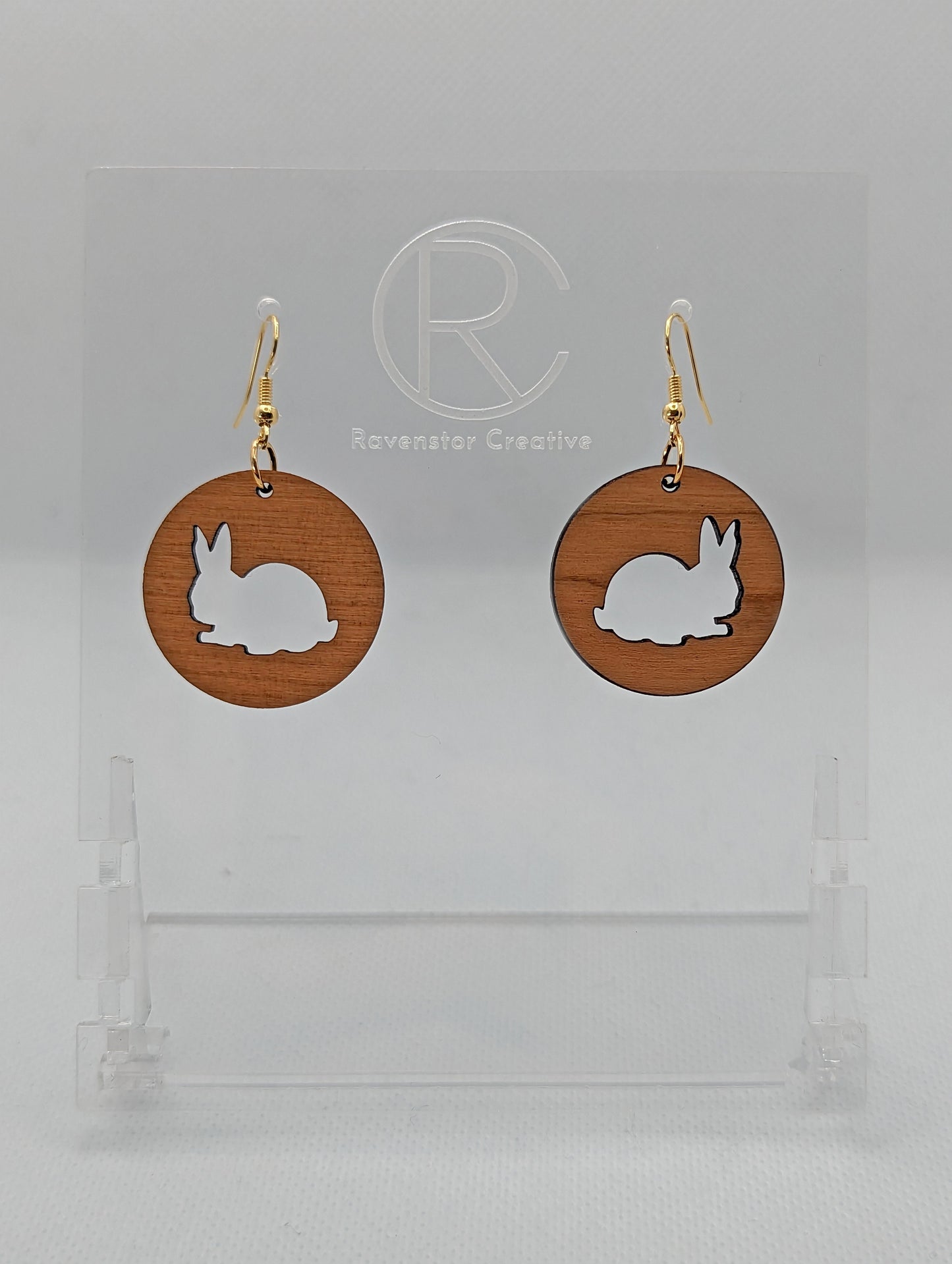 A pair of Wooden earrings that have Gold ear wire hook fixings. They are Circles with an Easter bunny / Rabbit cut out each one. It is made from Cherry Wood. They are against a white background on see through stands.
