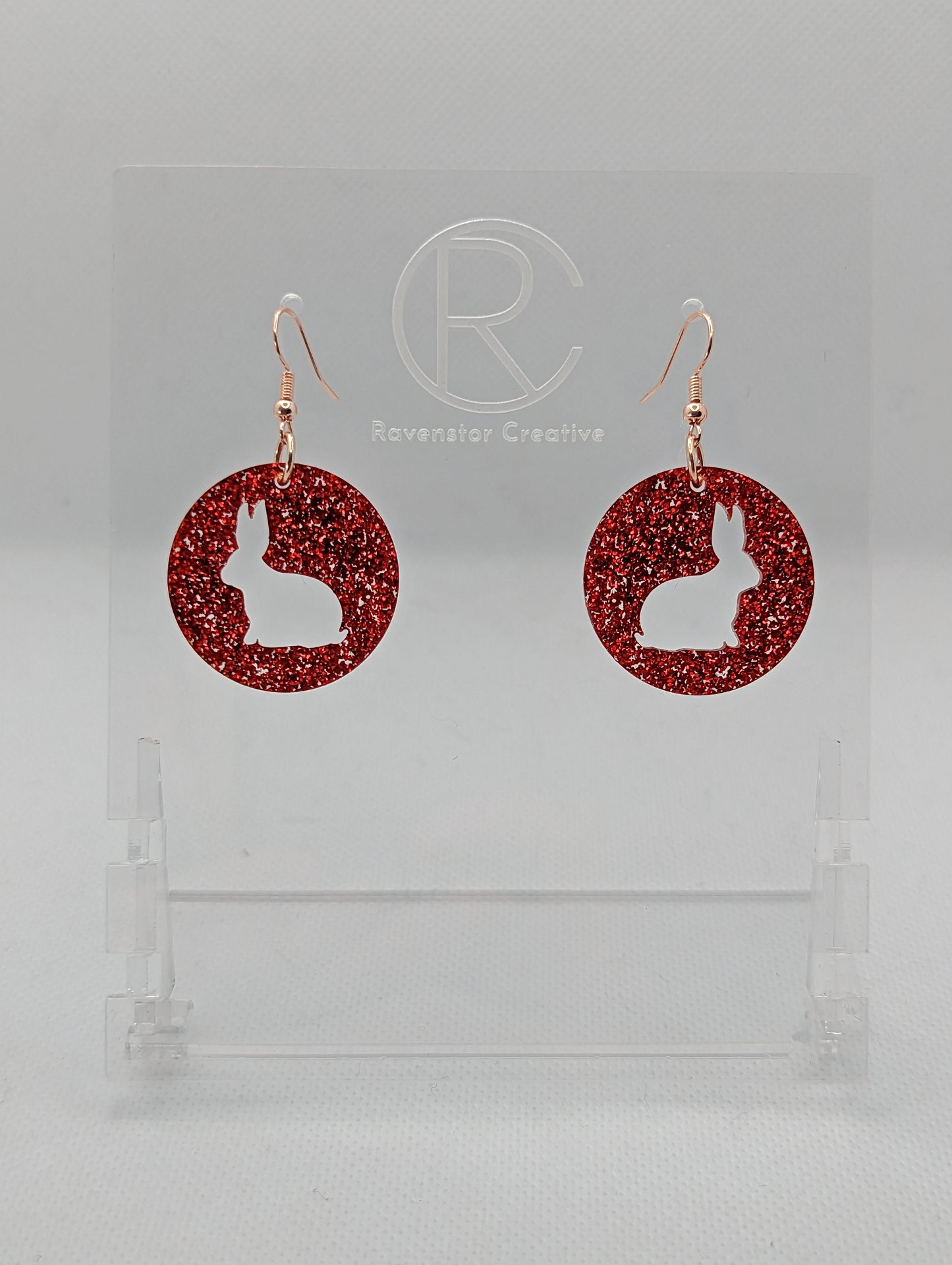 A pair of acrylic earrings that have Rose Gold ear wire hook fixings. They are Circles with an Easter bunny / Rabbit cut out each one. It is Red Glitter in colour. They are against a white background on see through stands.