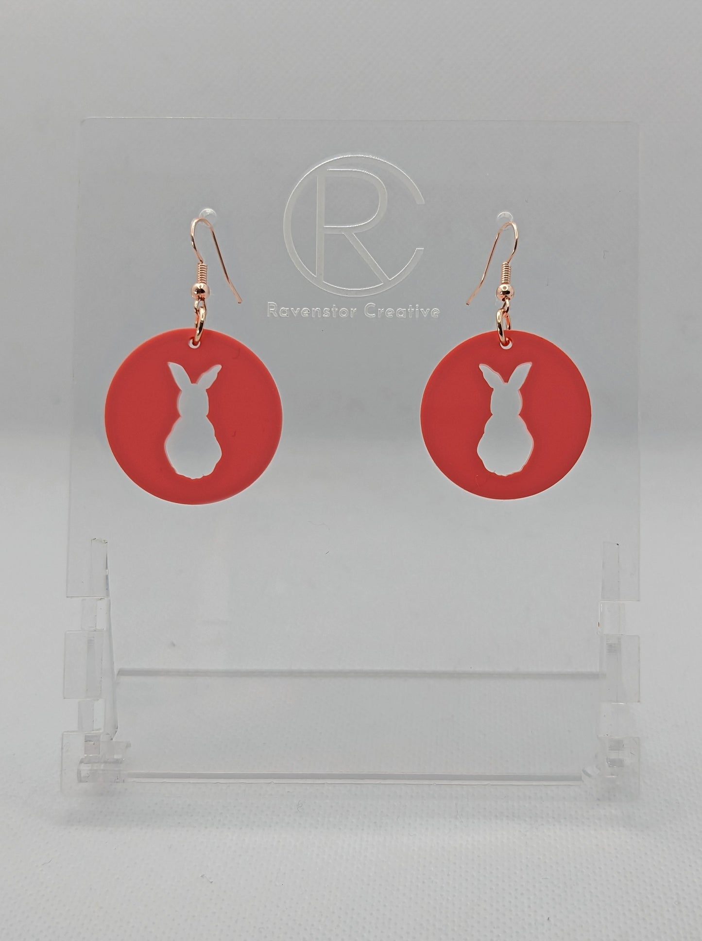 A pair of acrylic earrings that have Rose Gold ear wire hook fixings. They are Circles with an Easter bunny / Rabbit cut out each one. It is Sherbet Pink in colour. They are against a white background on see through stands.