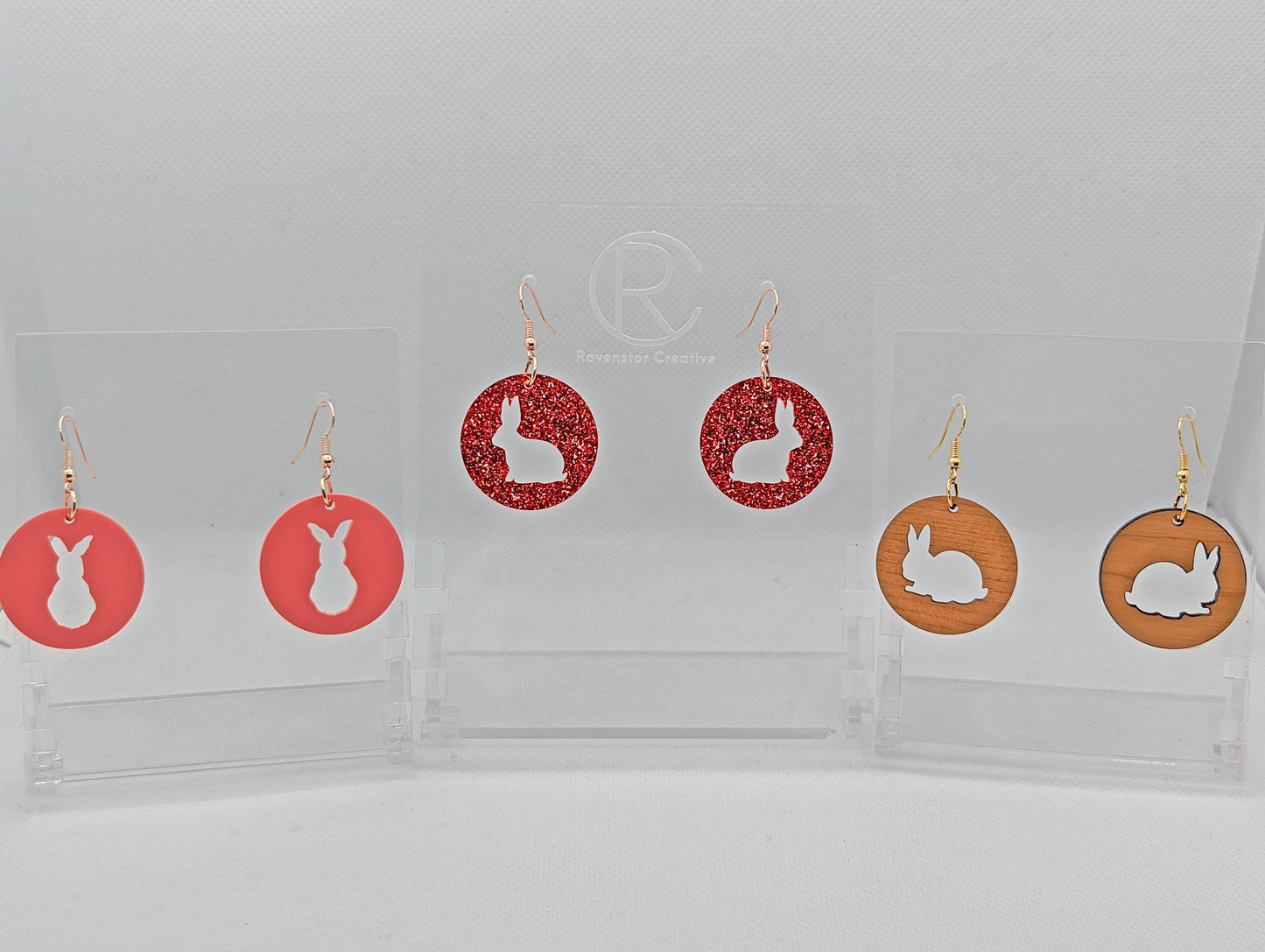 Tree pairs of earrings that have ear wire hook fixings. They are Circles with an Easter Rabbit / bunny cut out each one. The colours are Sherbet Pink and Red Glitter Acrylic and Cherry Wood. They are against a white background on see through stands.