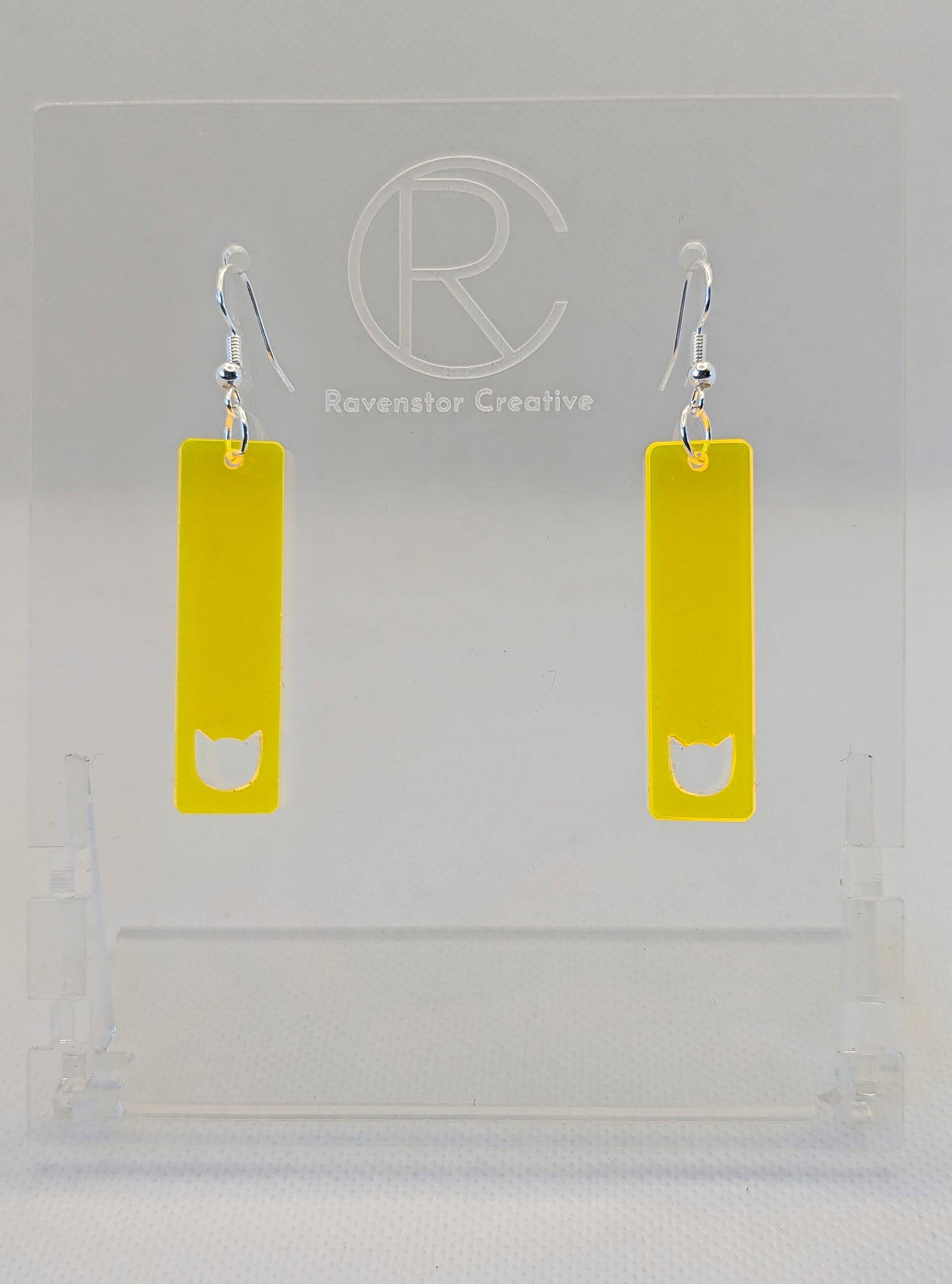 Shows a pair of acrylic earrings that have silver ear wire hook fixings. They are long and slim cat pendant earrings. They are neon yellow in colour and are against a white background on see through stand with the Ravenstor Creative logo on it.