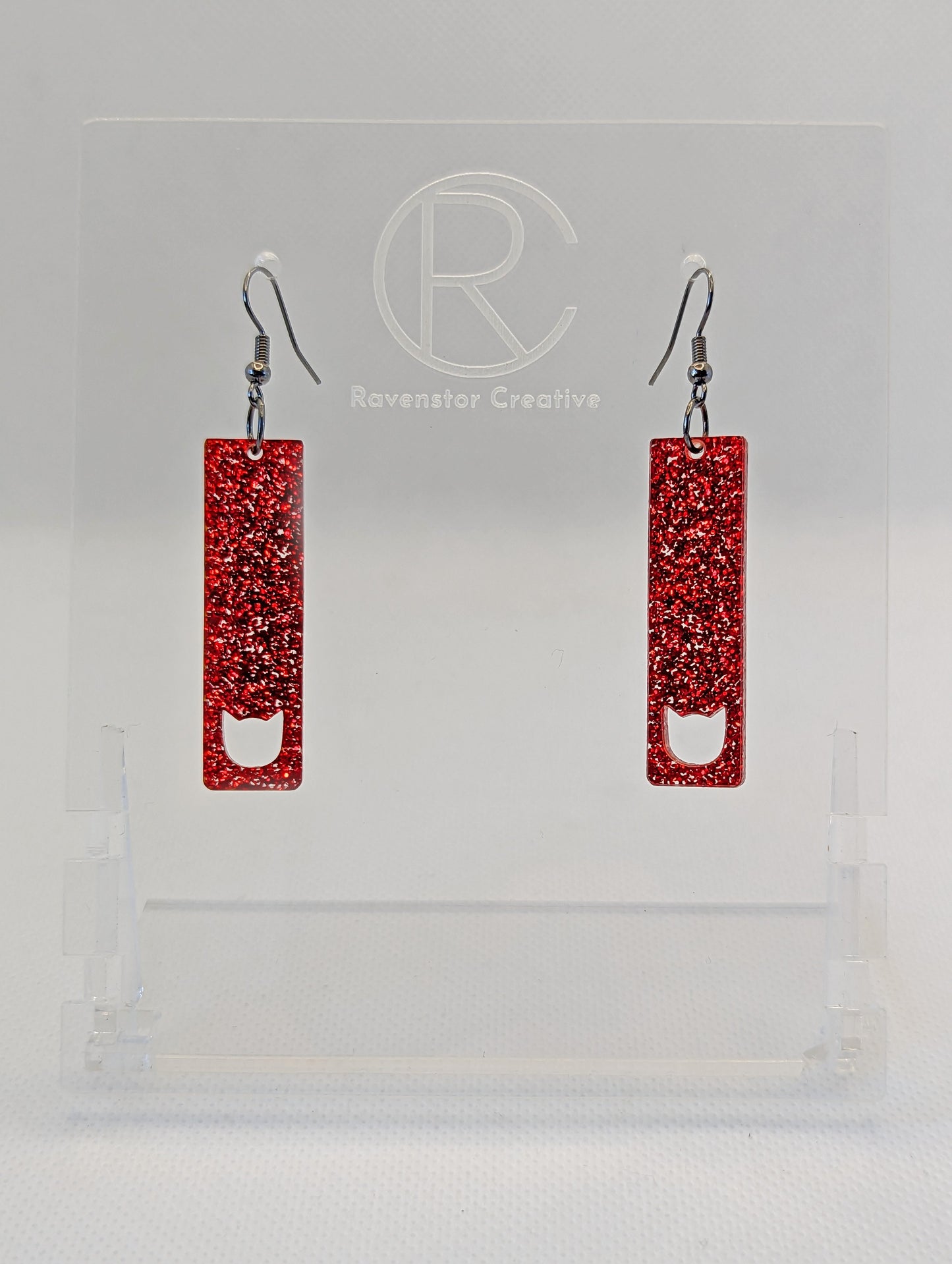 Image of a pair of slim cat pendant earrings on a hand showing the size against a white background, they are two and a half finger widths length and one wide. They are neon orange and have silver fixings.
