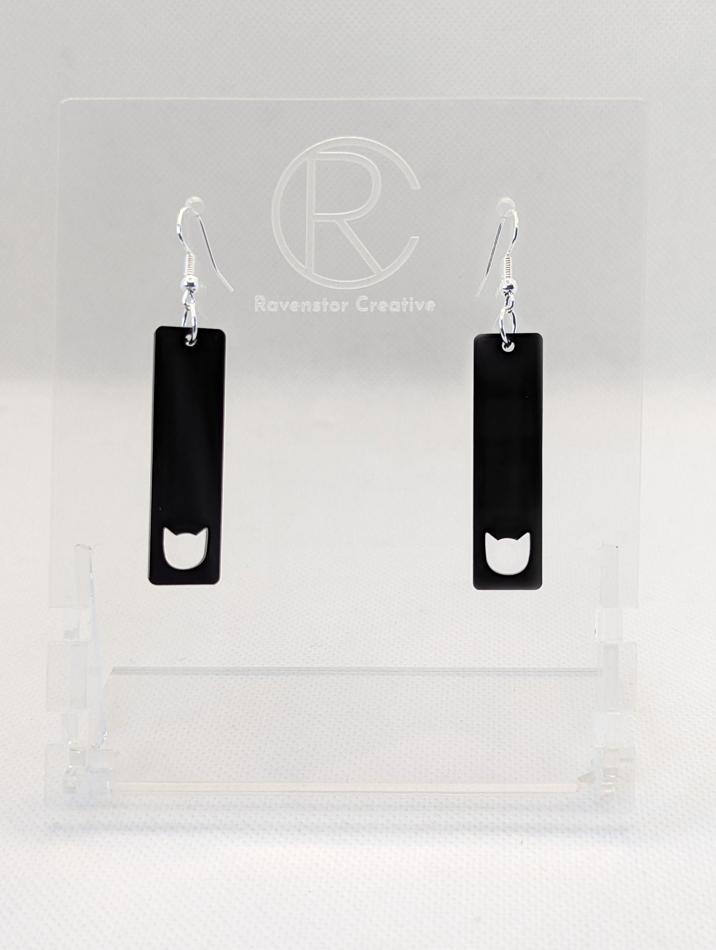 Shows a pair of acrylic earrings that have silver ear wire hook fixings. They are long and slim cat pendant earrings. They are black in colour and are against a white background on see through stand with the Ravenstor Creative logo on it.