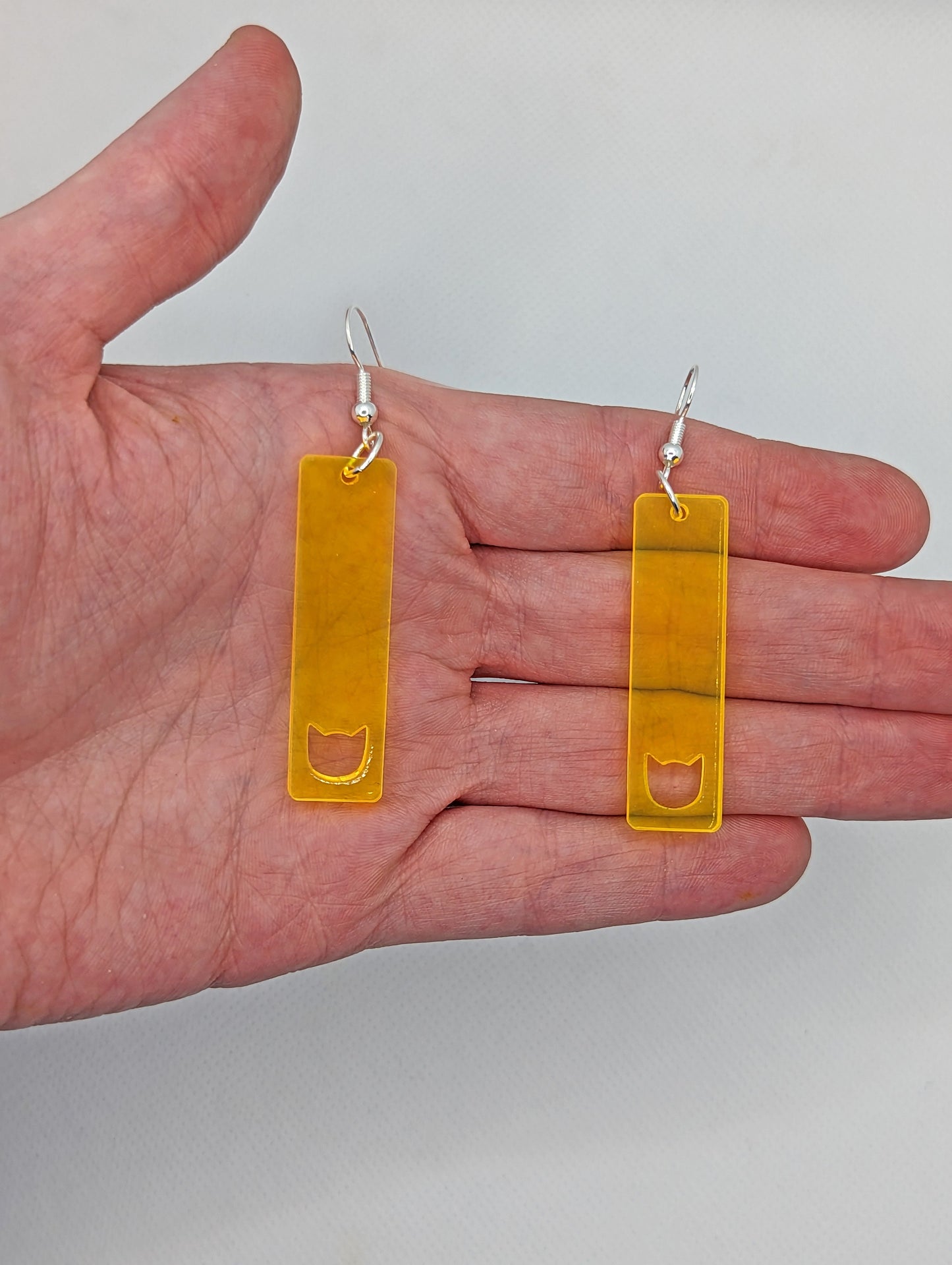 Image of a pair of slim cat pendant earrings on a hand showing the size against a white background, they are two and a half finger widths length and one wide. They are neon orange and have silver fixings.