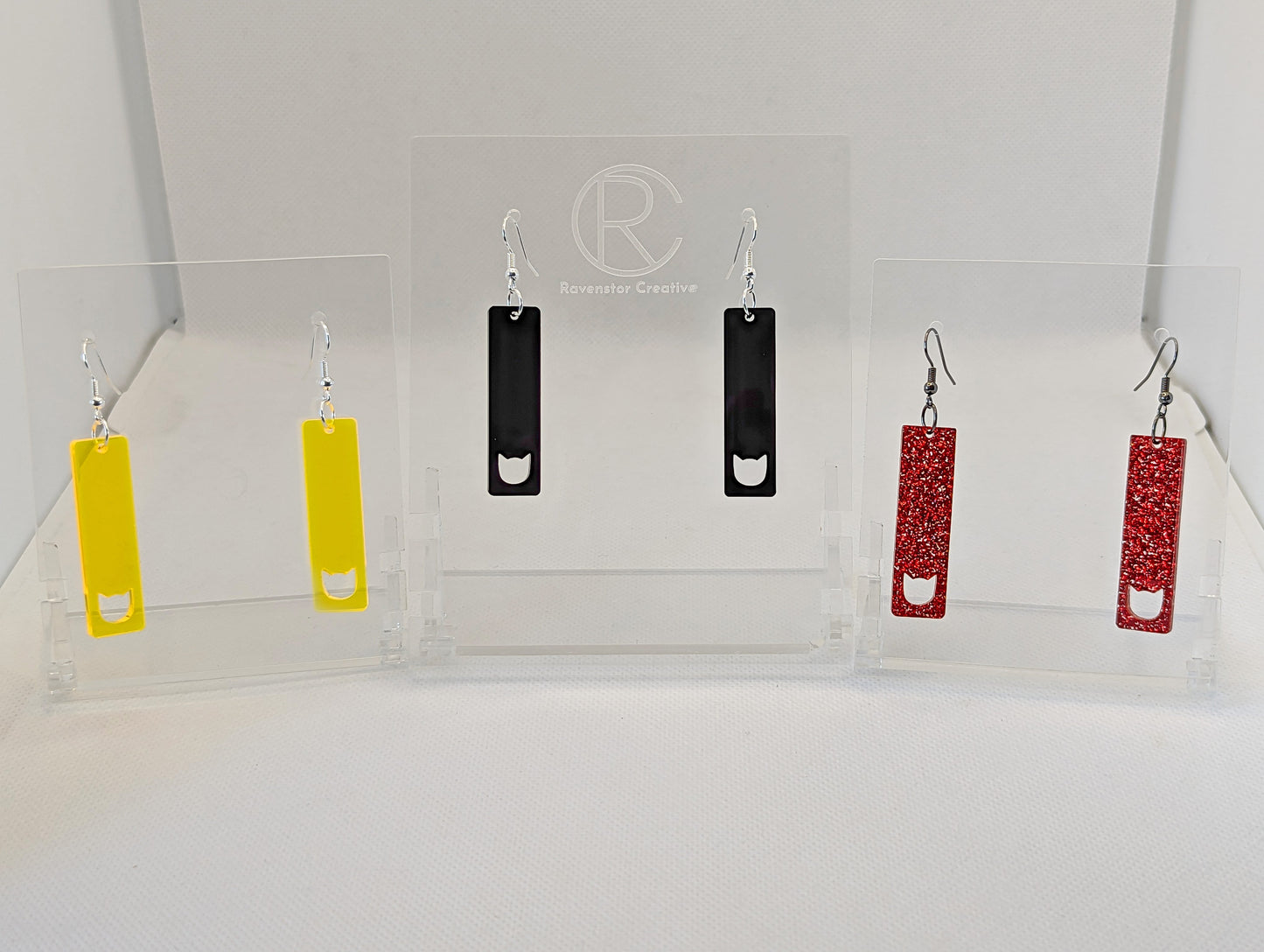Shows three pairs of earrings that have ear wire hook fixings. They are slim cat pendant earrings. The colours are neon yellow, black and red glitter. They are against a white background on see through stand with the ravenstor creative logo on.