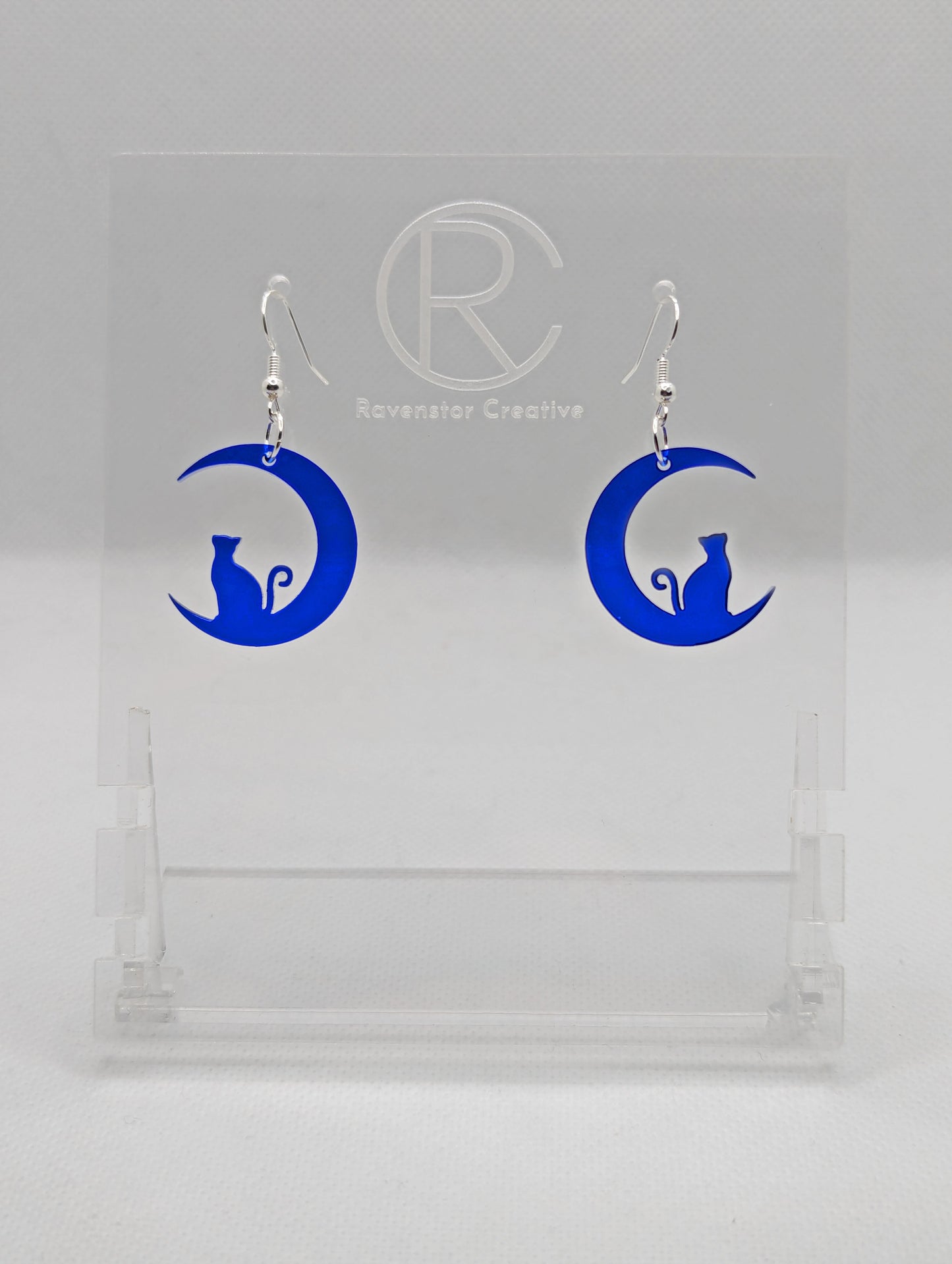 Shows a pair of acrylic earrings that have silver ear wire hook fixings. They are a cat sitting in a crescent moon. They are in translucent blue and are against a white background on see through stand with the Ravenstor Creative logo on it.