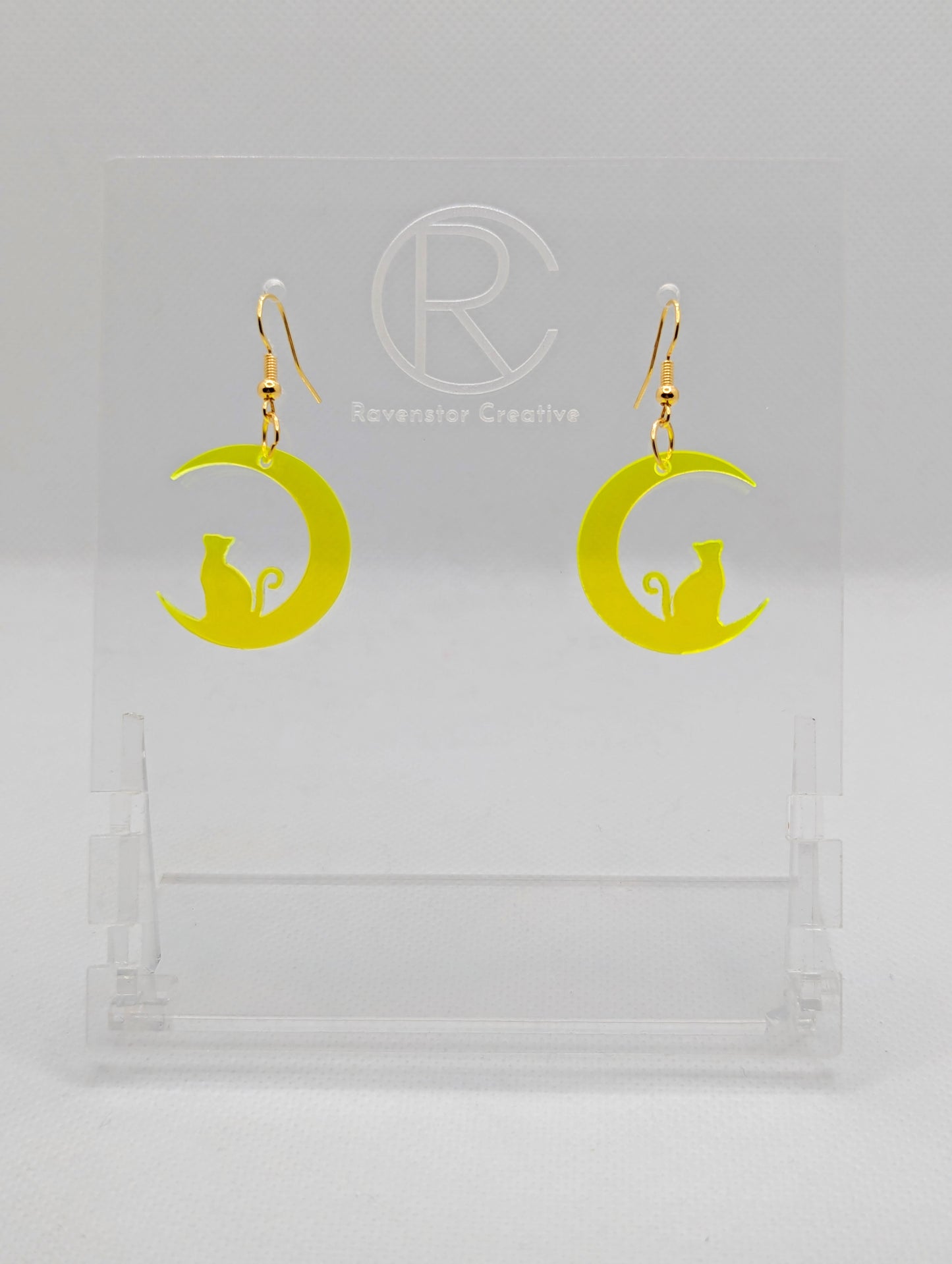 Shows a pair of acrylic earrings that have gold ear wire hook fixings. They are a cat sitting in a crescent moon. They are in neon yellow and are against a white background on see through stand with the Ravenstor Creative logo on it.