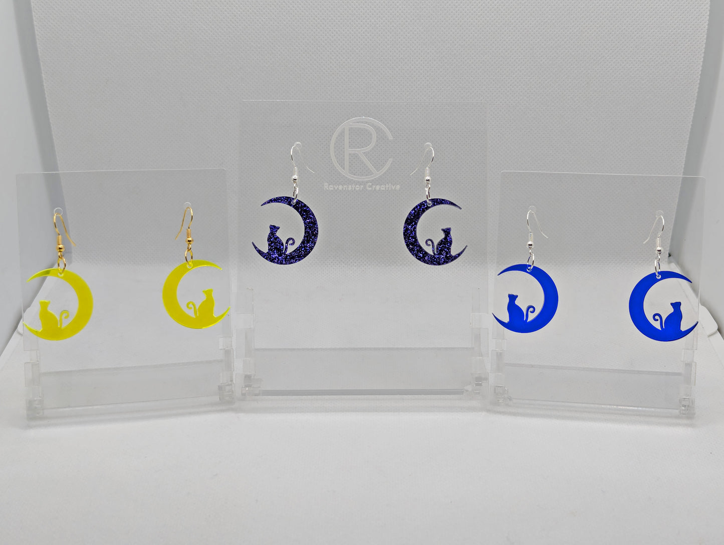 Shows three pairs of earrings that have ear wire hook fixings. They are a cat sitting on a crescent moon. The colours are Neon Yellow, Purple Glitter and Translucent Blue. They are against a white background on see through stands.