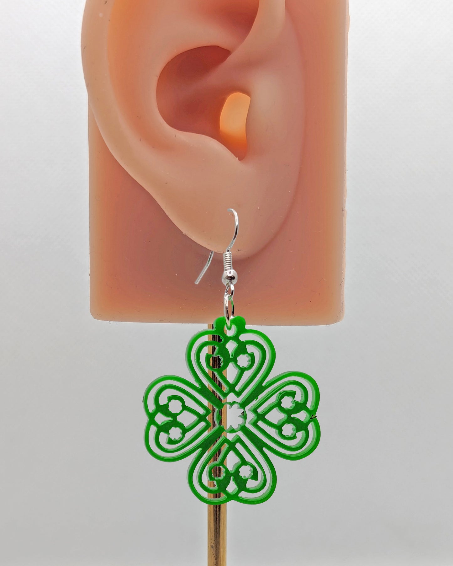 Shows an acrylic earring that have silver ear wire hook fixings. It is a four leaf clover design with Celtic patterns. It is on an ear showing the size against a white background.