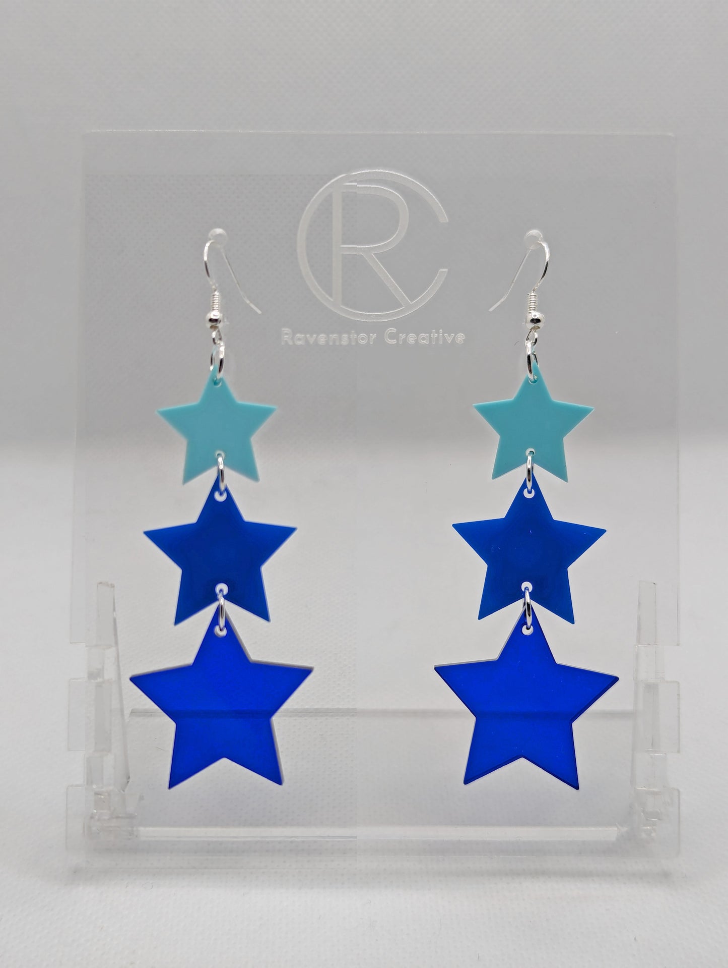 A pair of acrylic earrings with silver ear wire hook fixings. Each has three stars dangling from each other, the top is the smallest and they get bigger as they go down. The colours from the top are spearmint, blue and translucent blue.