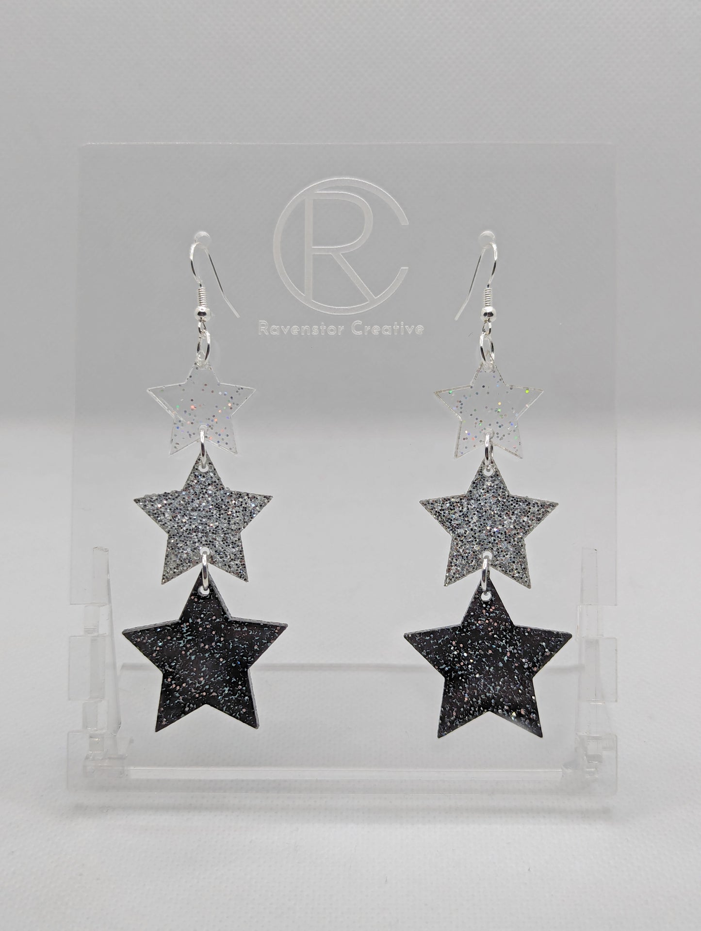 A pair of acrylic earrings with silver ear wire hook fixings. Each has three stars dangling from each other, the top is the smallest and they get bigger as they go down. The colours from the top are clear sparkle, silver glitter and black glitter.