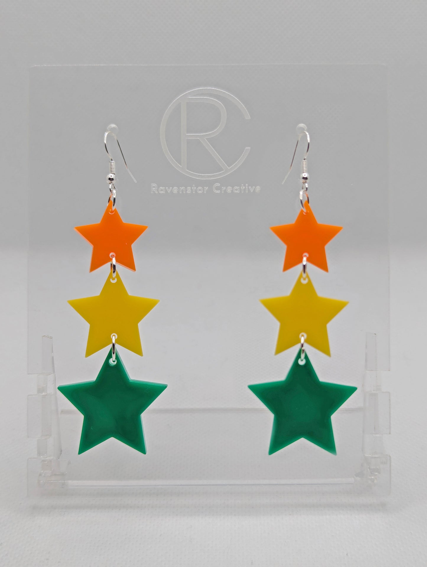 A pair of acrylic earrings with silver ear wire hook fixings. Each has three stars dangling from each other, the top is the smallest and they get bigger as they go down. The colours from the top are orange, yellow and green.