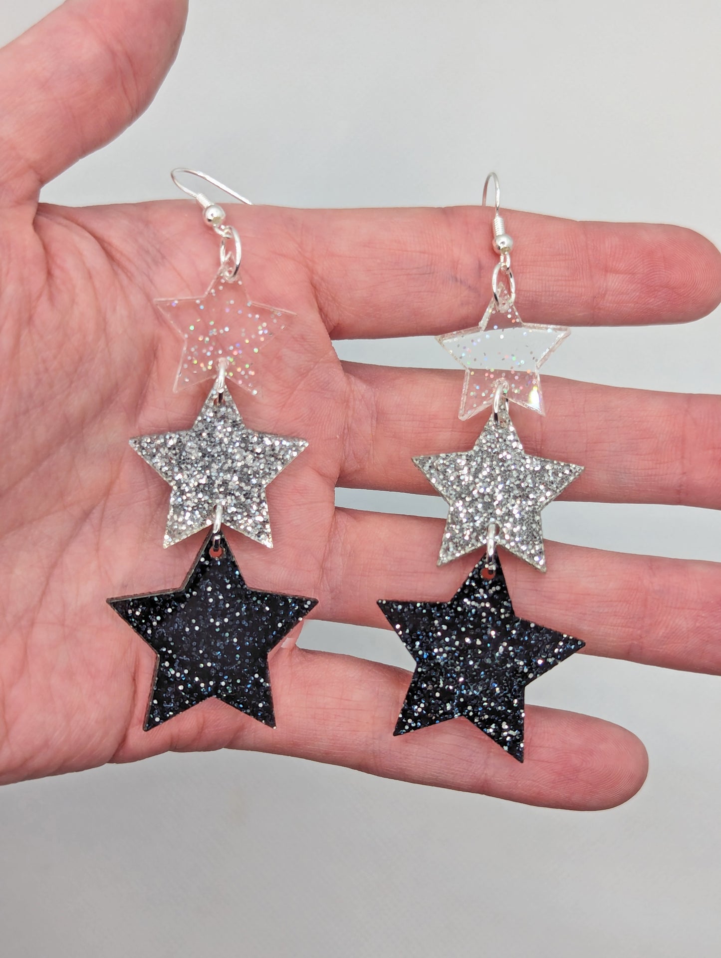 A pair of dangly earrings, each has three stars dangling from each other, the top is the smallest and they get bigger as they go down. They are against a hand to show the size. They are four finger widths in length and the bigger heart is two wide.