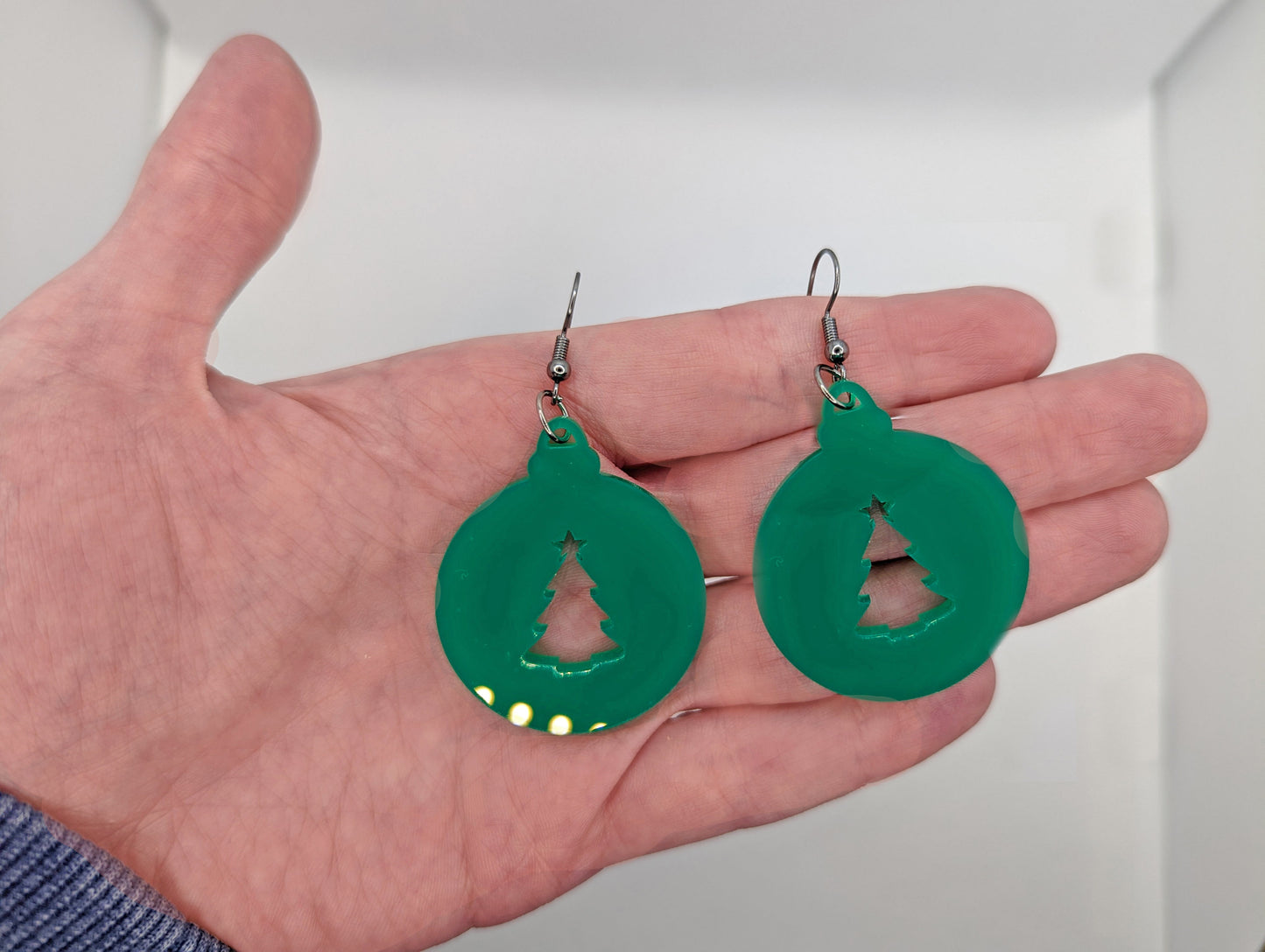 Image of a pair of earrings. They are Christmas bauble shapes with an Xmas tree cut out in Green on a hand showing the size against a white background, they are 2 finger widths length the same again wide. They and have silver fixings.
