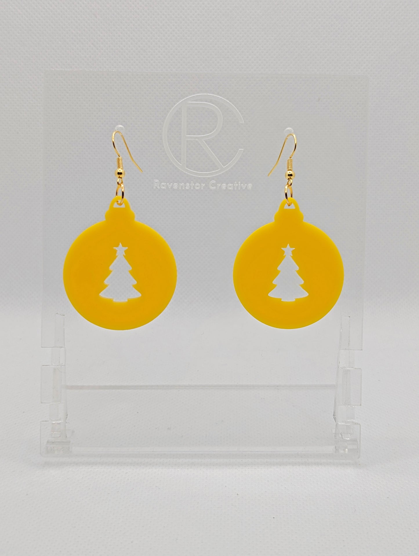 Shows a pair of acrylic earrings that have rose gold ear wire hook fixings. They are Christmas baubles shapes with an Xmas tree shape cut out the middle. The colour is Yellow. They are against a white background on see through stands.