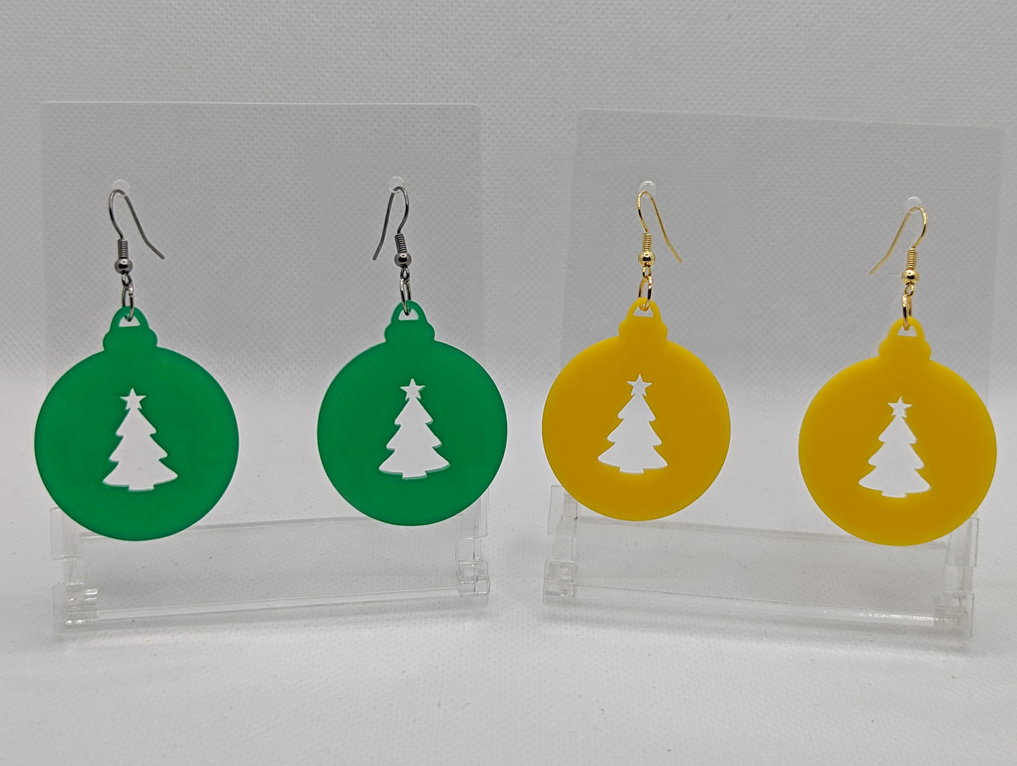 Shows two pairs of earrings that have ear wire hook fixings. They are Christmas baubles with an Xmas Tree shape cut out the middle. The colours are Green and Yellow. They are against a white background on see through stands.