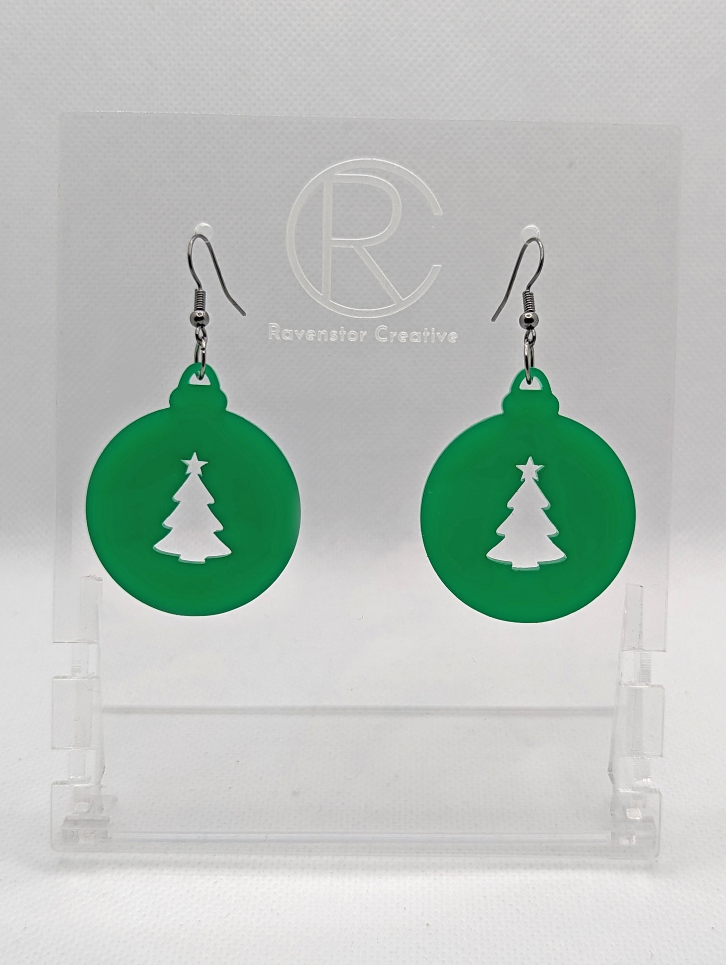 Shows a pair of acrylic earrings that have black ear wire hook fixings. They are Christmas baubles shapes with an Xmas tree shape cut out the middle. The colour is Green. They are against a white background on see through stands.