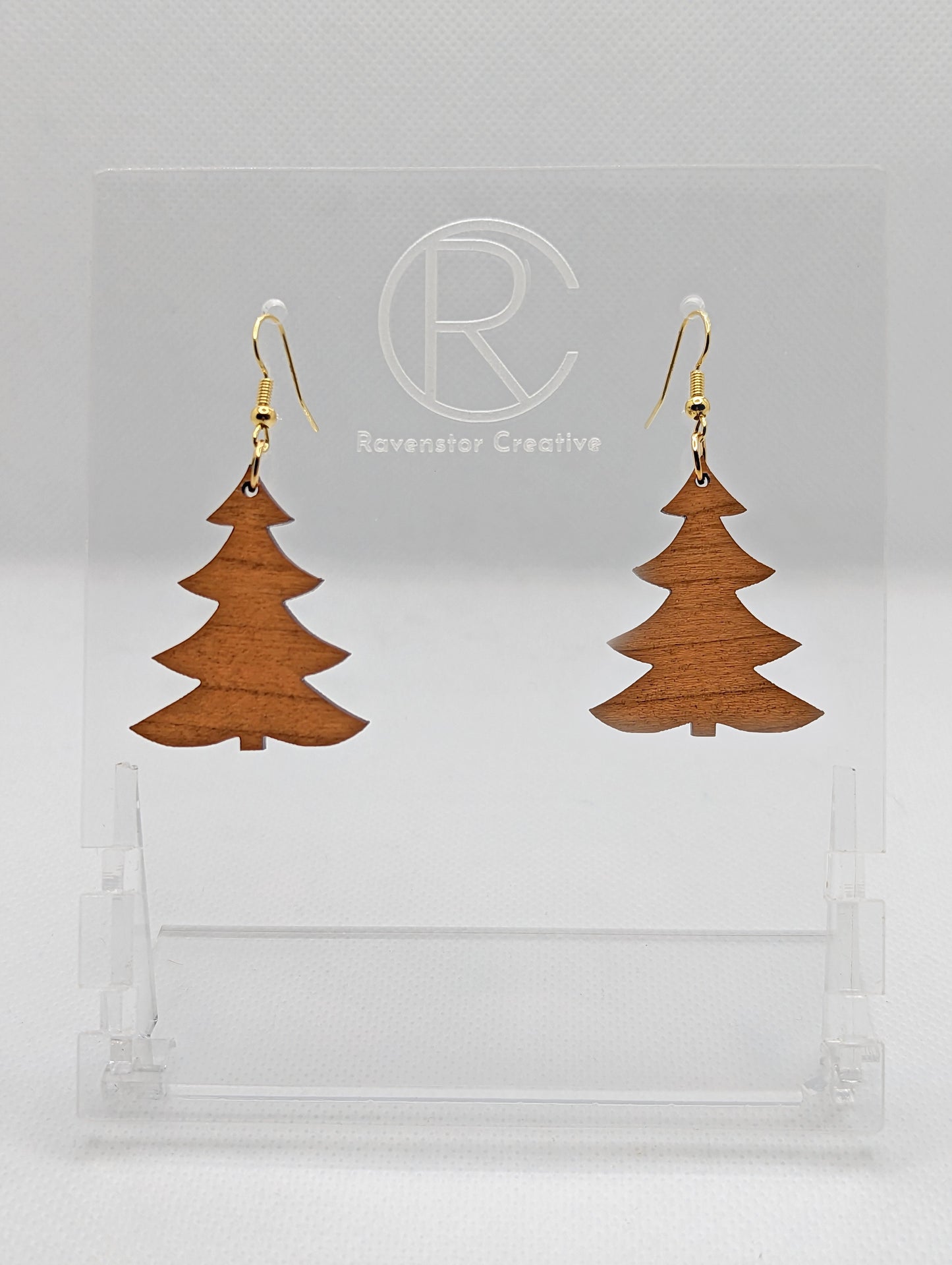 An image of a pair of earrings with Gold ear wire hook fixings. They are Christmas / Xmas Tree shapes. They are made in Cherry Wood. They are dangling from a clear stand with the Ravenstor Creative logo on it against a white background