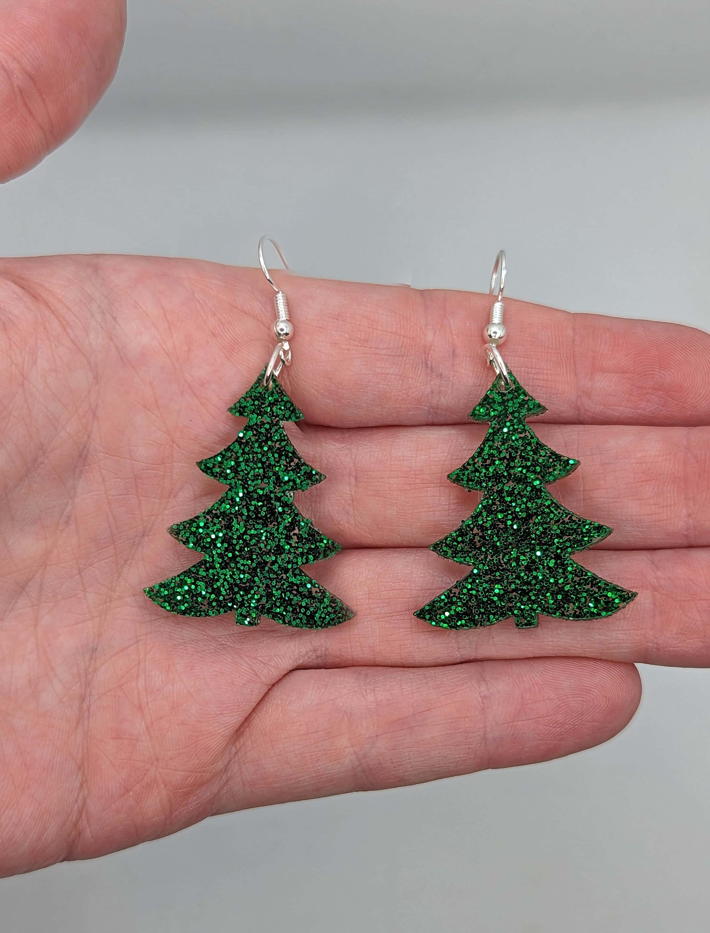 An Image showing a pair of Christmas Tree earrings in Green glitter with silver fixings. They are against a hand to show the size of Two and a half finger widths in length and two wide. They are against a white background.