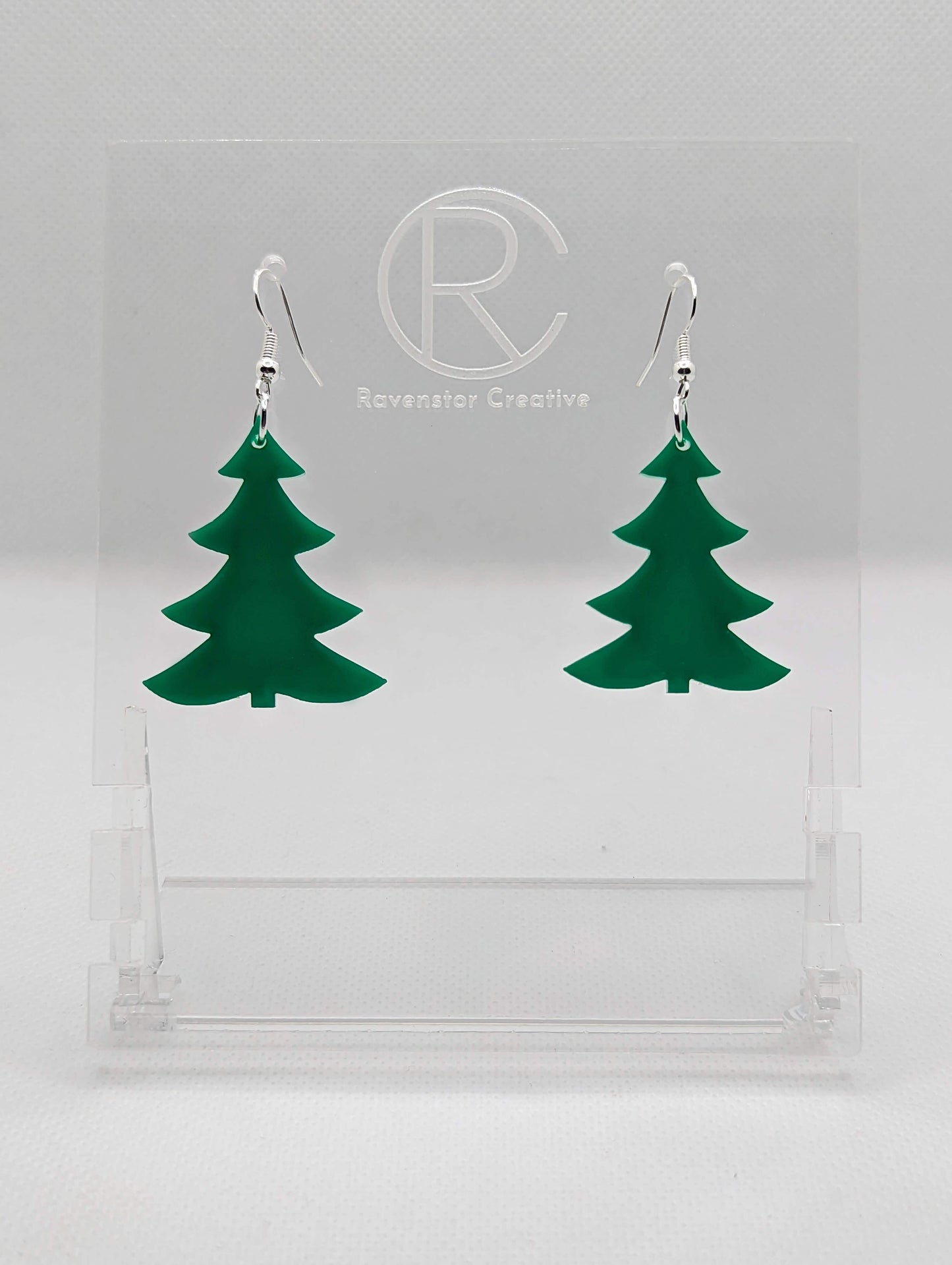 An image of a pair of earrings with Silver ear wire hook fixings. They are Christmas / Xmas Tree shapes. They are made in Green acrylic. They are dangling from a clear stand with the Ravenstor Creative logo on it against a white background