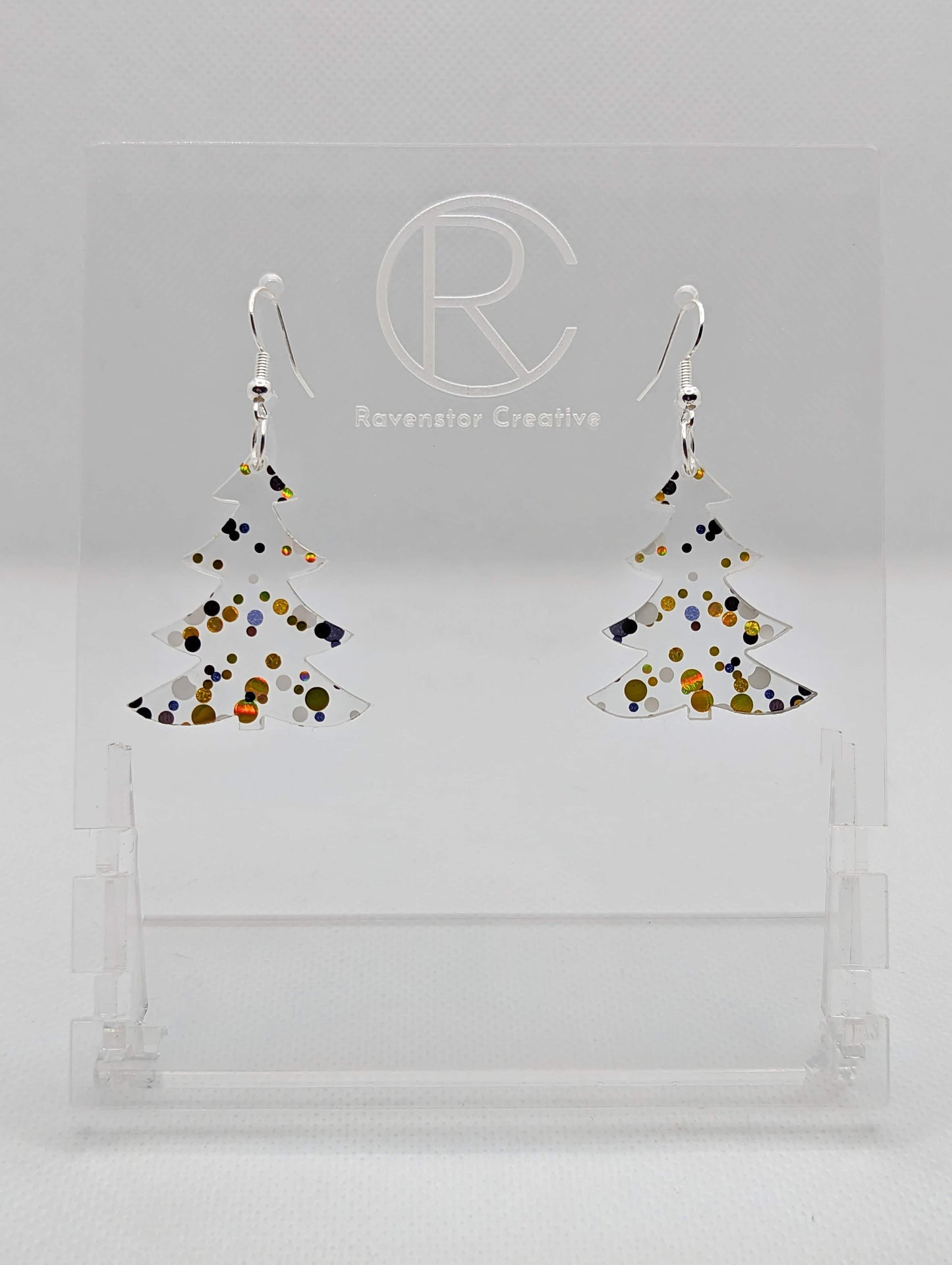 An image of a pair of earrings with Silver ear wire hook fixings. They are Christmas / Xmas Tree shapes. They are made in Lollipop Glitter acrylic. They are dangling from a clear stand with the Ravenstor Creative logo on it against a white background