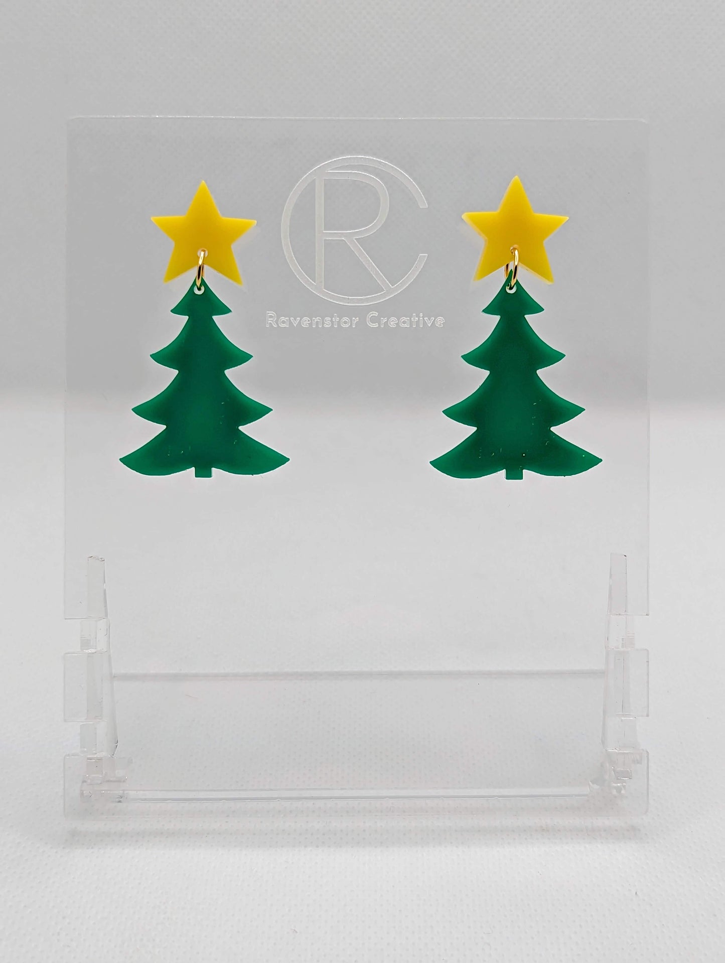 A pair of acrylic Christmas tree earrings in two parts that have stud fixings. The studs are attached to a star with the tree dangling under They have a Yellow star with a Green Christmas Tree Dangling underneath. They are displayed on a clear stand.