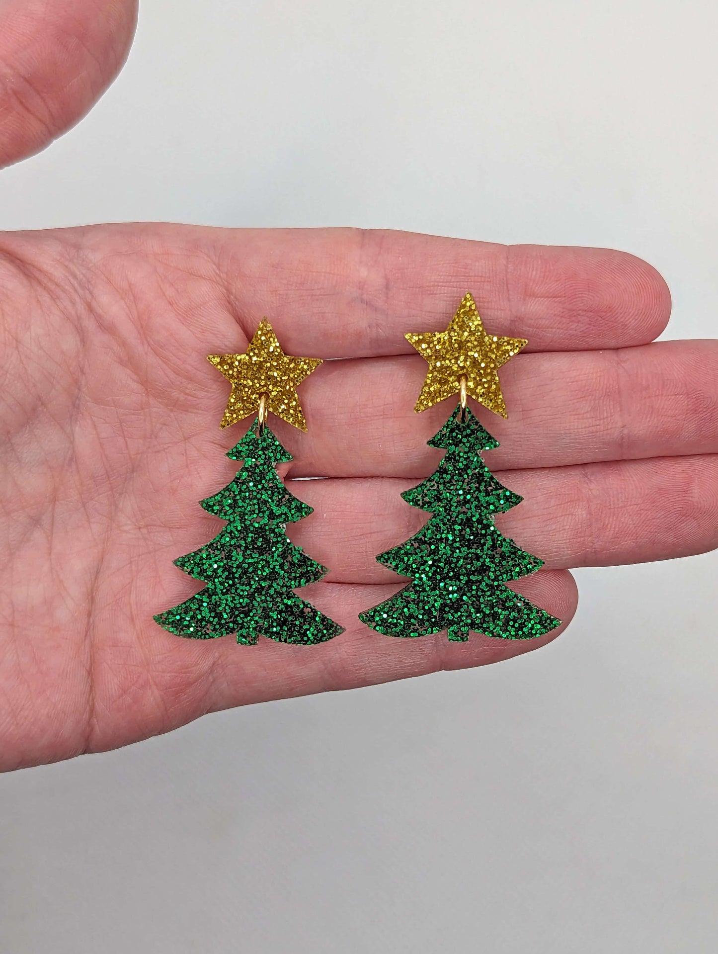 A pair of Christmas tree earrings in two parts that have stud fixings. The stud are attached to a star with the tree dangling under. They are against a hand to show the size of three finger widths in length and two wide. Colour Gold and green glitter