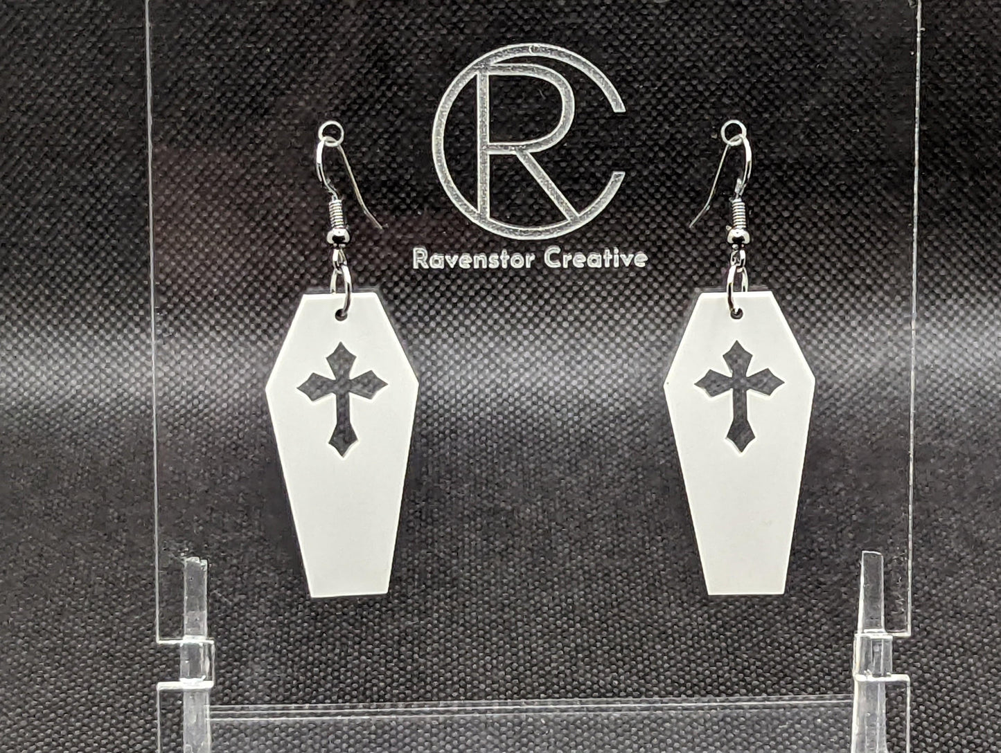 Shows a pair of earrings with black ear wire hook fixings. They are Spooky Halloween coffins / Casket shapes. The colour of them is white. They are on a see through stand that has the Ravenstor Creative Logo and its against a dark background.