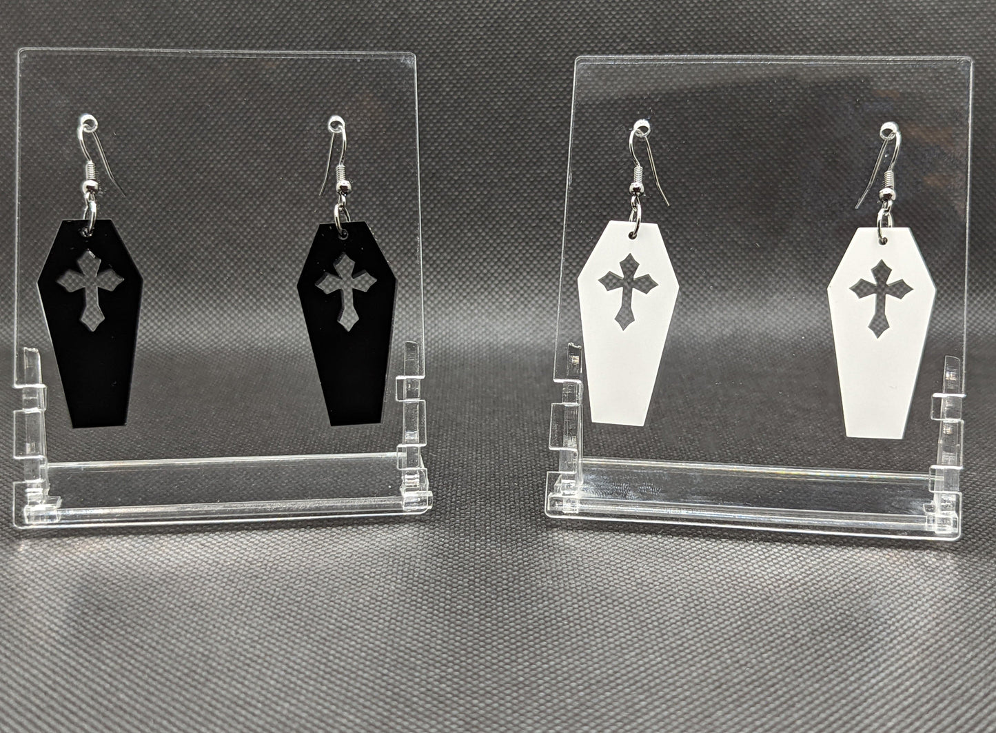 Shows 2 pairs of silver earrings with ear wire hook fixings. They are Halloween Coffins / Casket shapes. The colours of them are black and white. They are on see through stands with the Ravenstor Creative logo on them against a dark background.